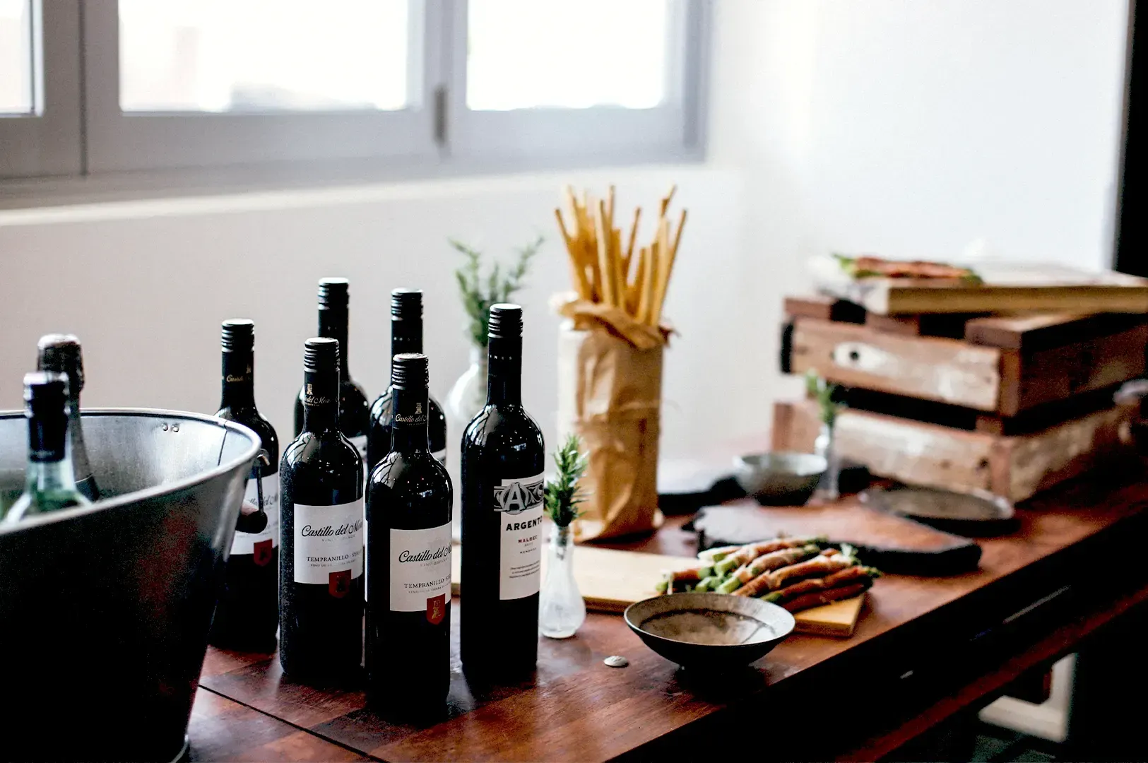 A rustic wooden table set with wine bottles, a metal chilling bucket, grissini breadsticks, and smal
