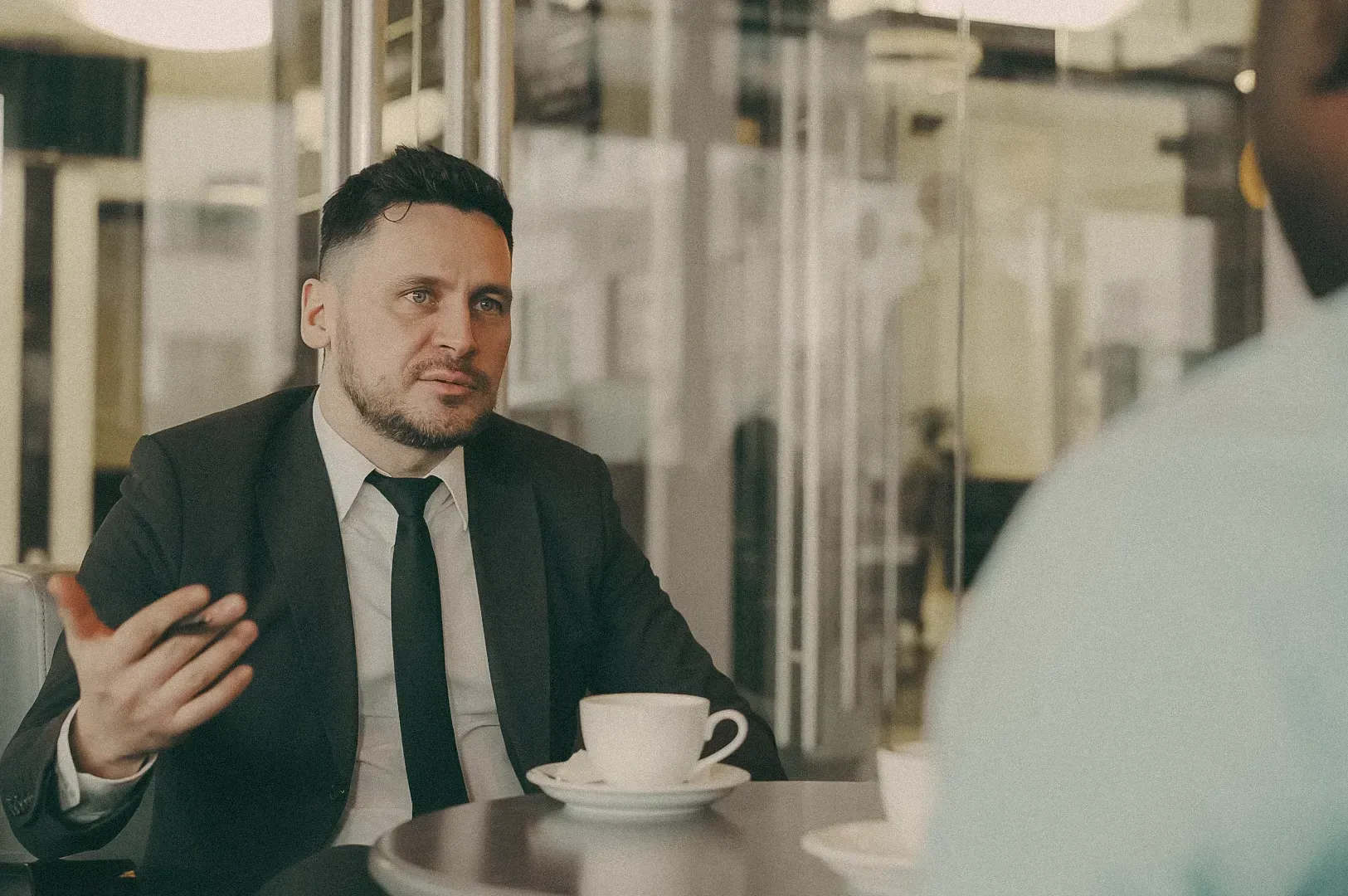 Professional in a suit speaking during a meeting at a café-style table with documents and coffee cup