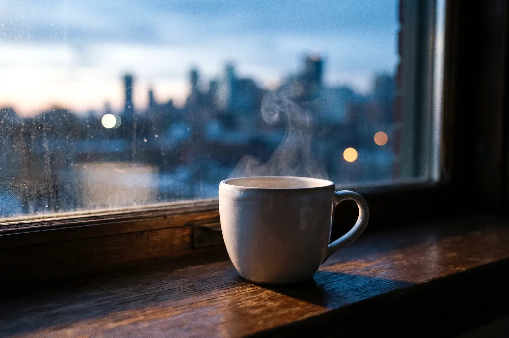 Close-up eye-level shot of a steaming coffee cup on a wooden windowsill overlooking a soft-focus cit