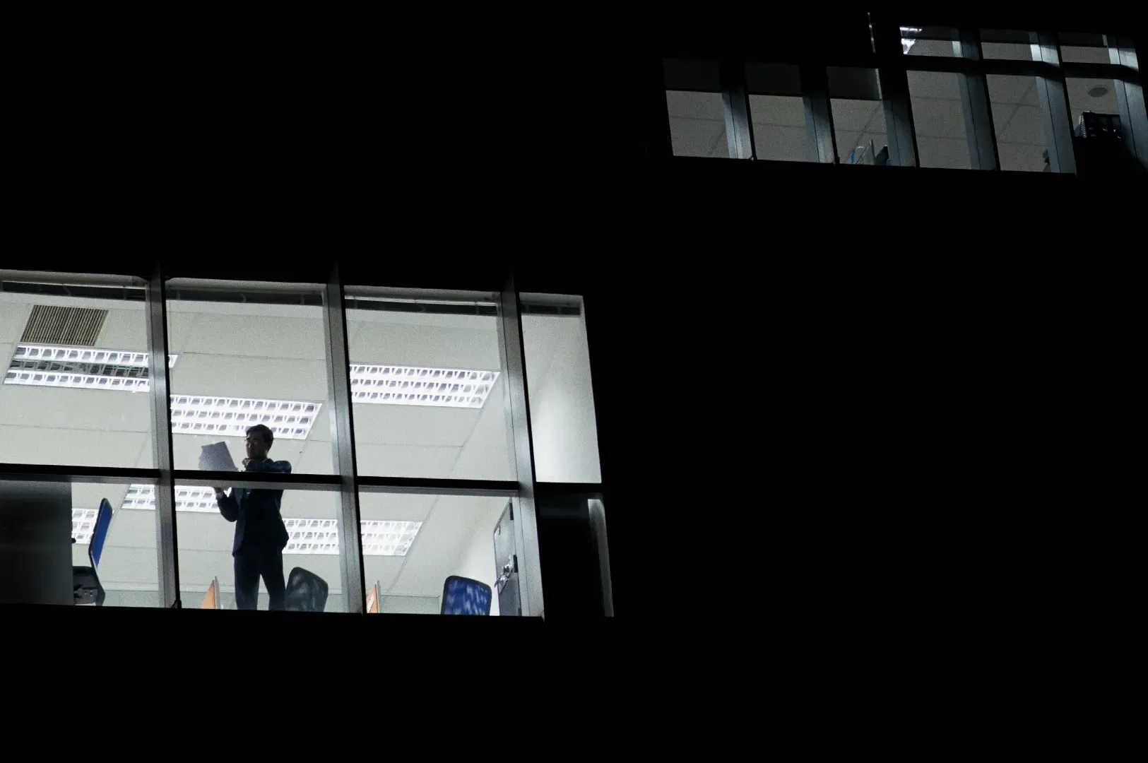 Exterior long shot of an office building at night showing a single professional standing inside a br