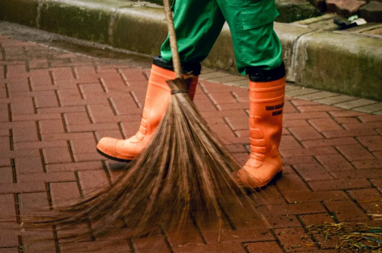 Low-angle motion shot of a street cleaner sweeping a wet brick sidewalk with a broom, capturing quie