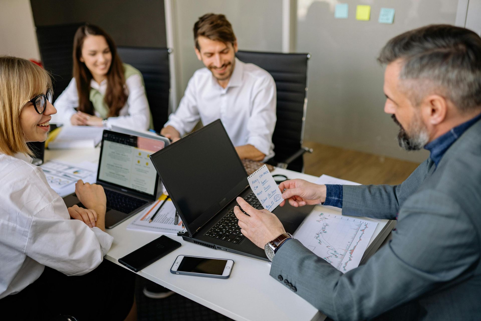 A group of four professionals are gathered around a white office table, engaged in a collaborative meeting with laptops, smartphones, and financial charts spread out before them. A man in a grey blazer holds up a small handwritten note to show his colleagues while everyone maintains a positive and focused atmosphere.