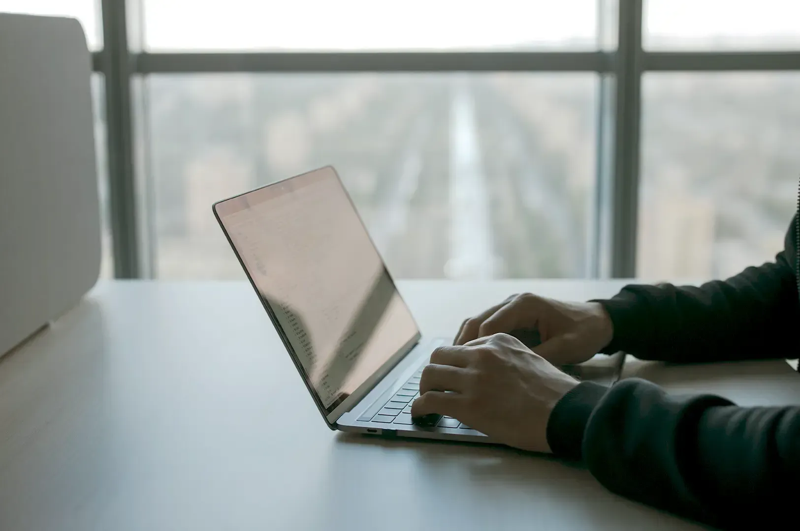 A close-up of hands typing on a laptop computer by a bright window, representing the small, consistent, and often unseen daily efforts that compound into long-term success.