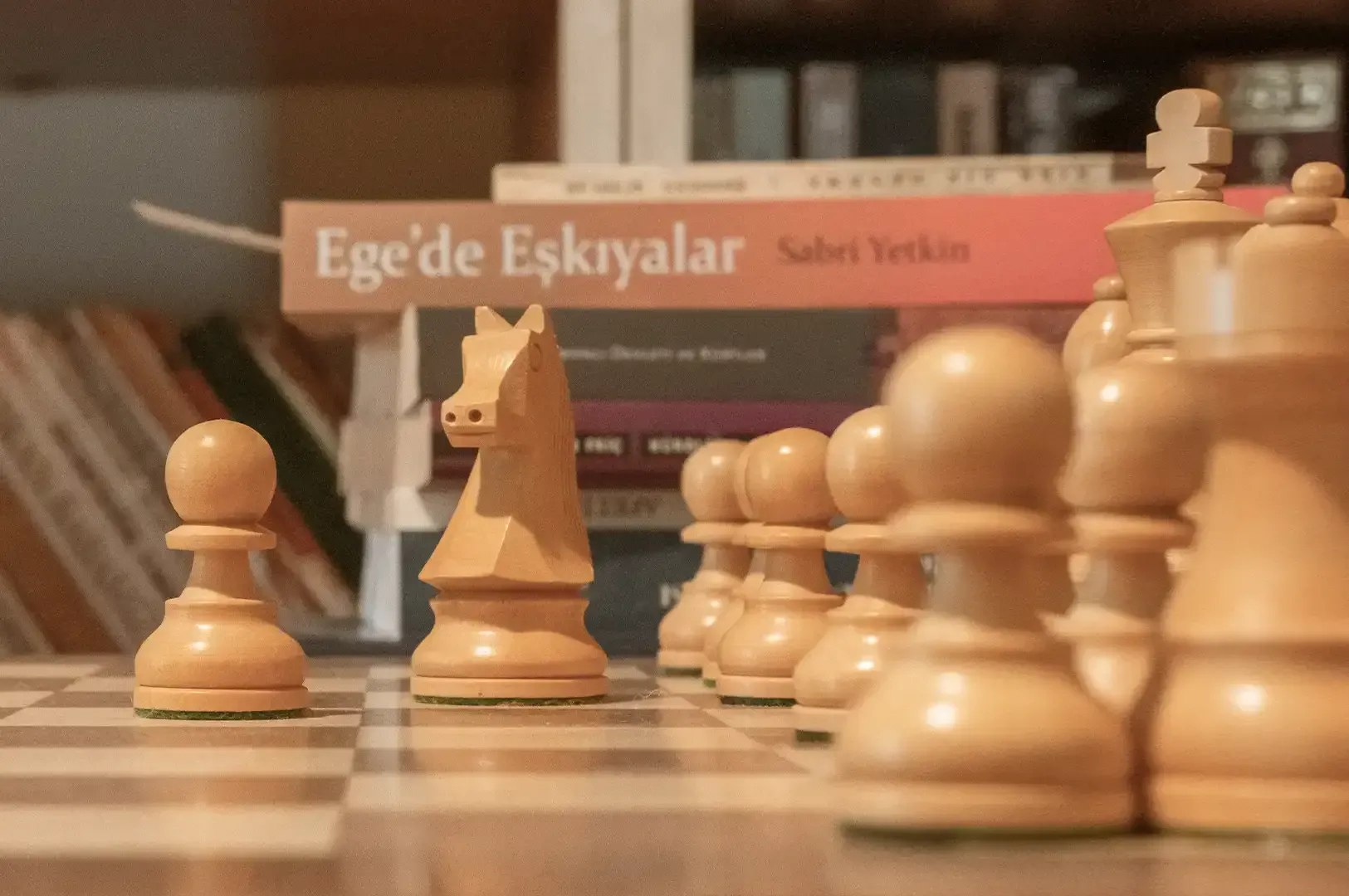 A close-up shot of classic wooden chess pieces lined up on a chessboard against a backdrop of stacked books, symbolizing the mindful attention, strategy, and deliberate focus championed in The Art of Noticing.