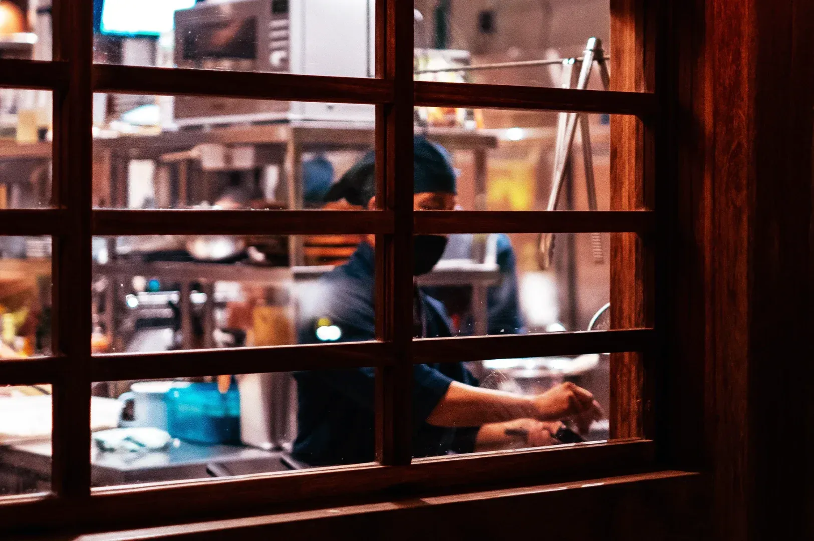 A view through a traditional wooden-latticed window of a culinary professional at work, representing the preservation of methods and the legacy of care in professional environments.