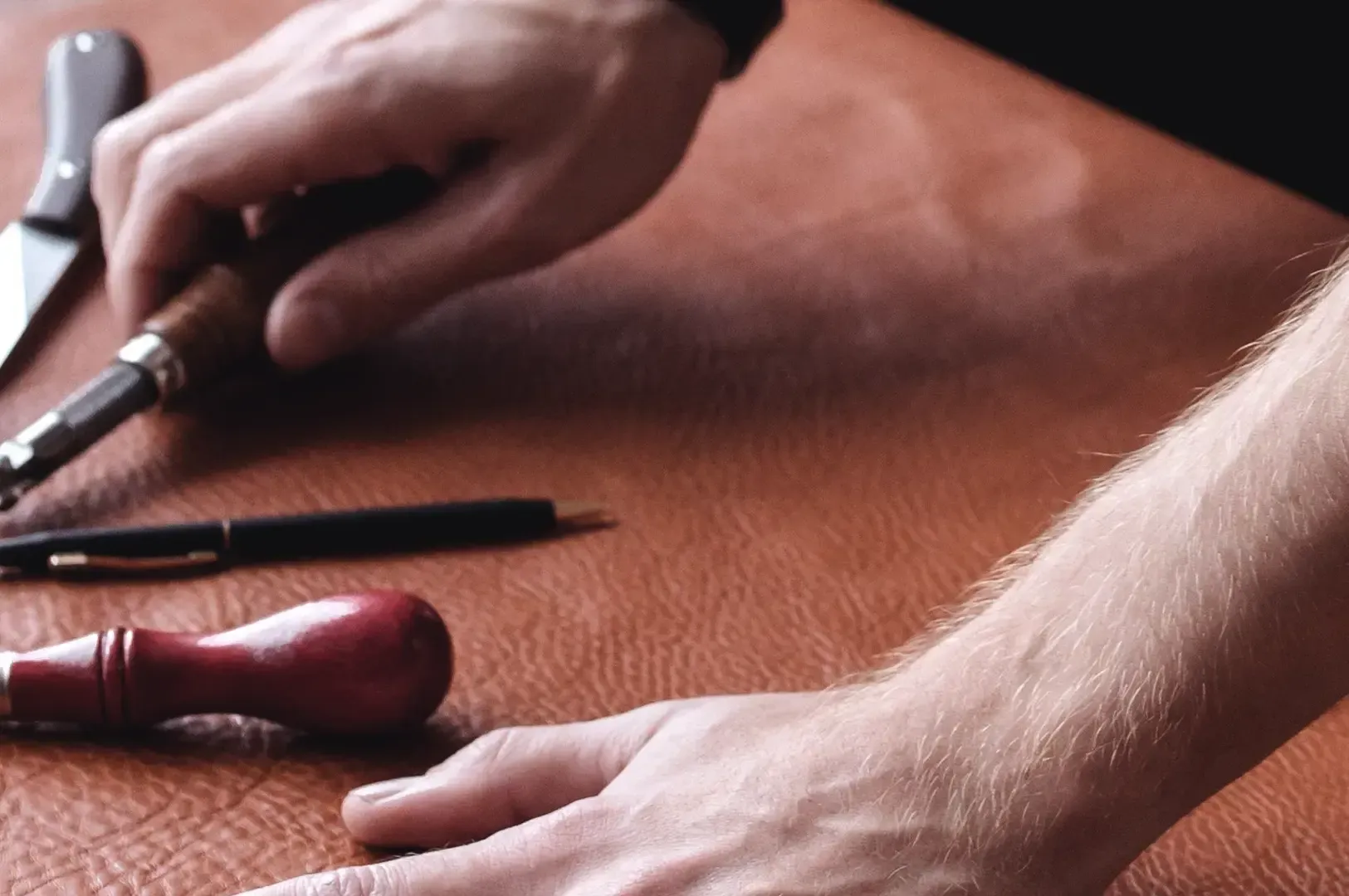 A craftsman's hands carefully smoothing a piece of rich brown leather alongside traditional leatherworking awls and blades, highlighting quality standards and physical judgment.