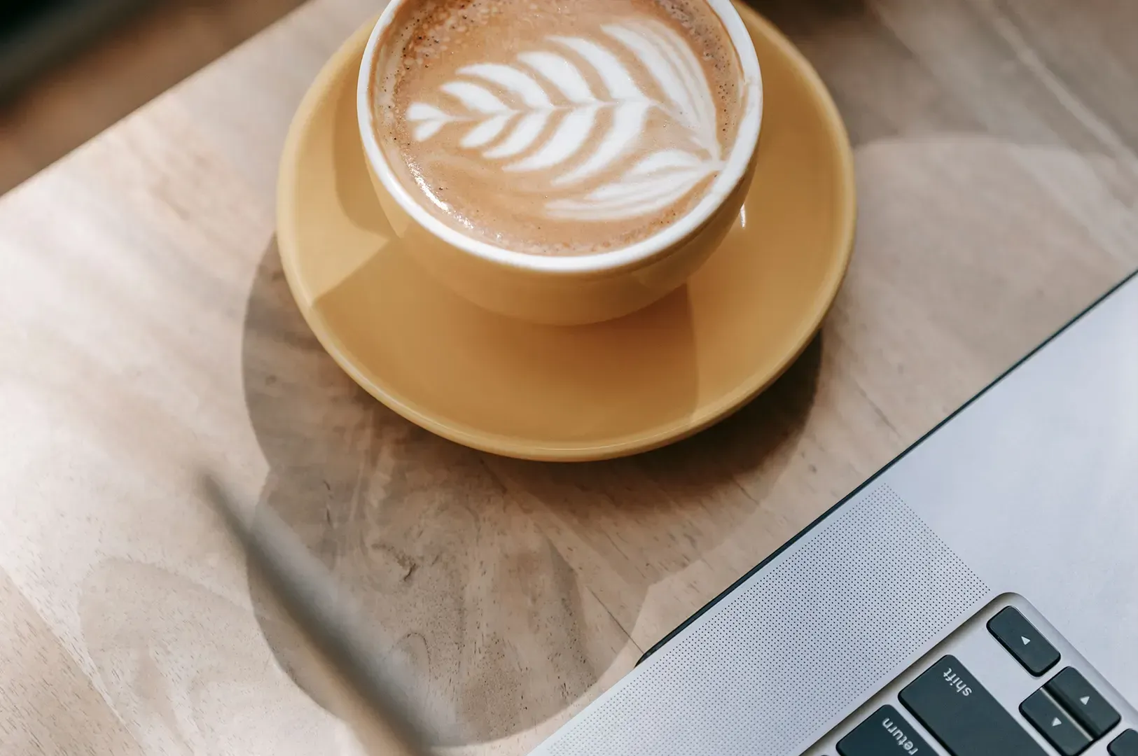 Top-down shot of a yellow ceramic cup of latte art coffee on a matching saucer next to a laptop keyboard on a wooden table, emphasizing slow observation, mindfulness, and building things that last by noticing.