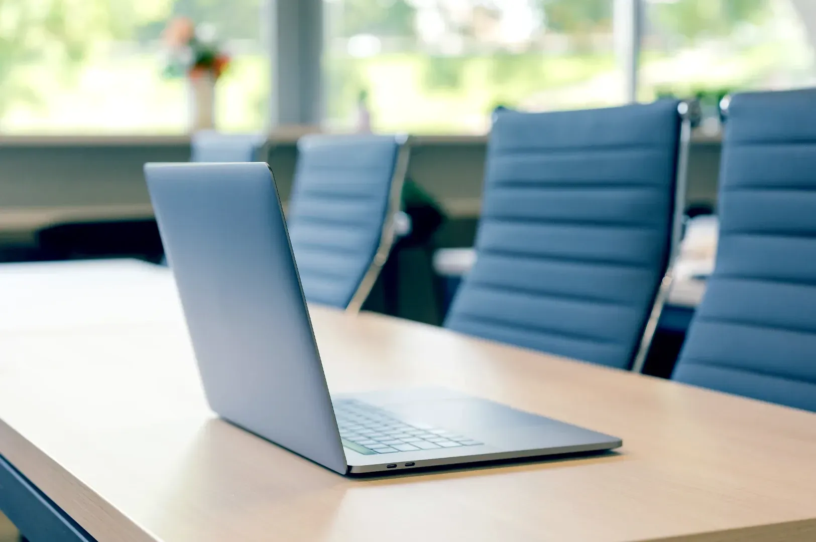 Close-up of an open laptop sitting on a clean wooden boardroom table with modern office seating in the background, symbolizing focused work environments.