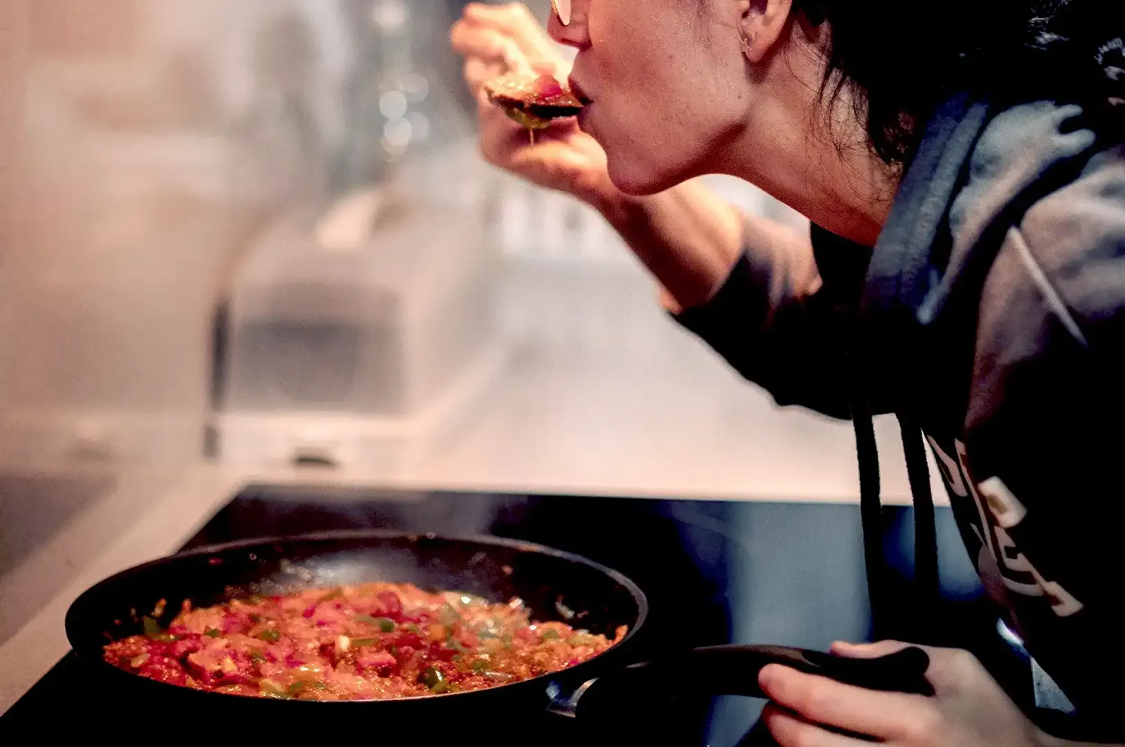 A home cook tasting food from a spoon while preparing a savory dish in a frying pan on a modern stovetop, reflecting the craft and identity explored in The Taste of Place.