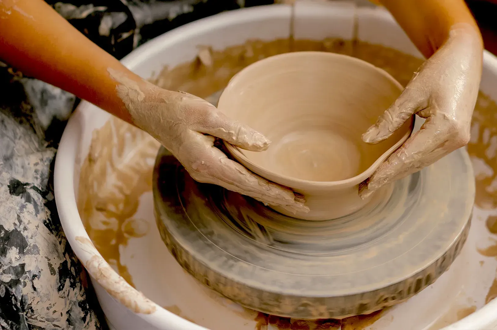 The hands of an artisan potter covered in clay, working on a bowl atop a spinning wheel, capturing the tactile and imperfect human process behind handmade craft.