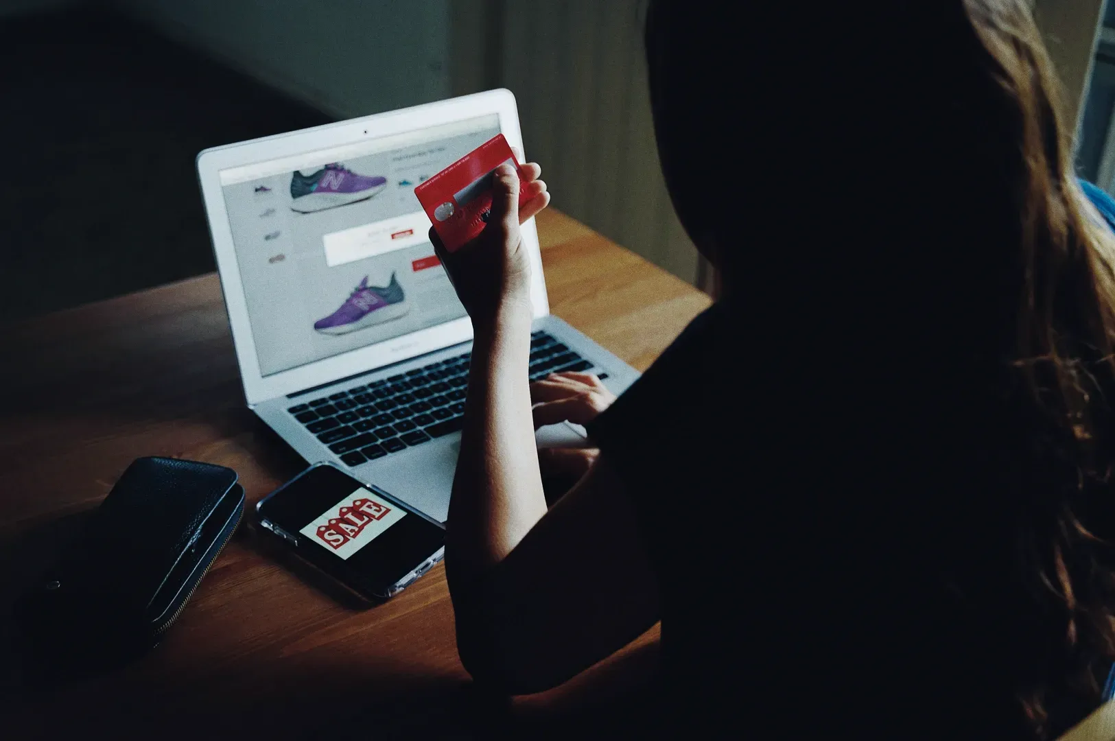 Over-the-shoulder medium shot of person shopping online holding a credit card beside a laptop and smartphone with sale sign, illustrating fast consumerism, impulse buying, and disposable culture contrasted with conscious consumption