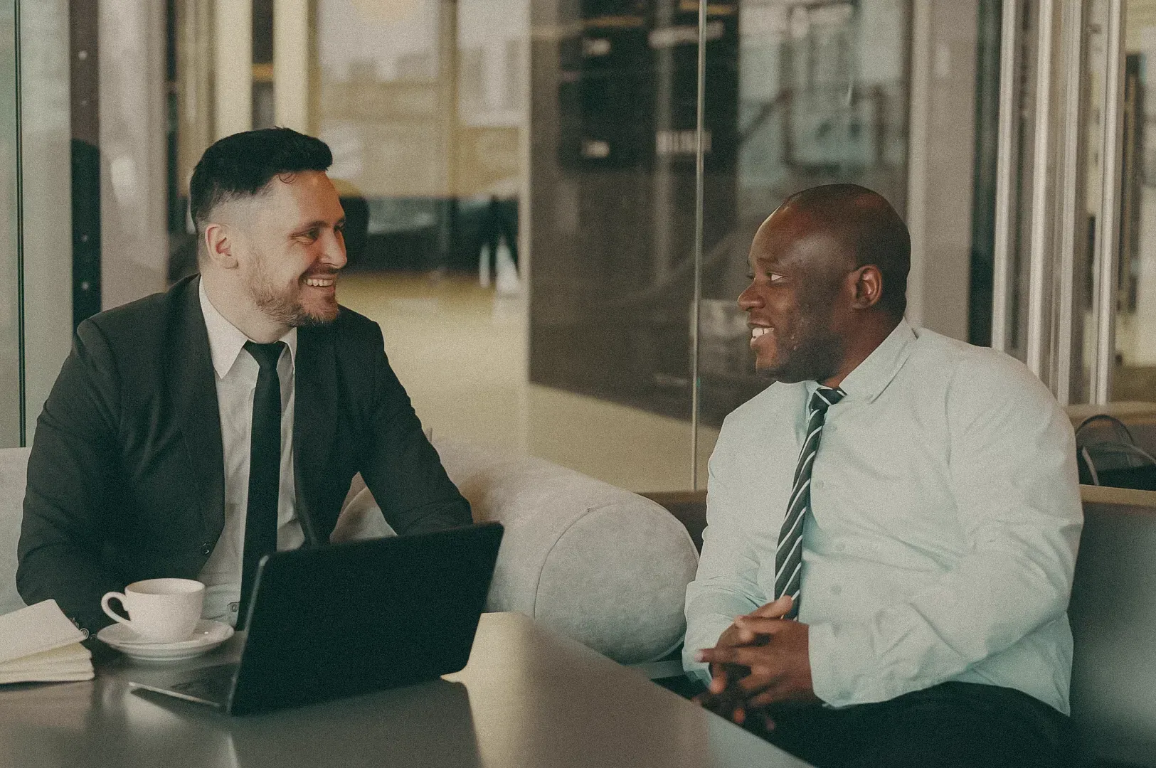 Two professionals having a in a modern office lounge, seated with a laptop and coffee on the table.