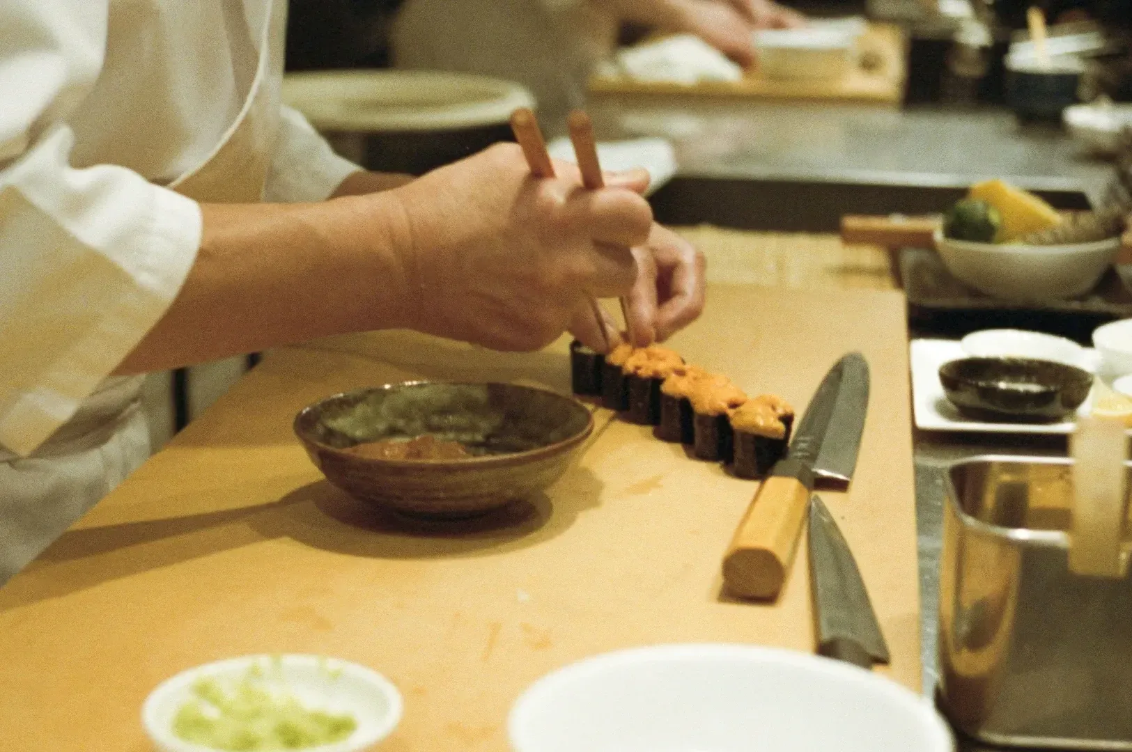 Close-up over-the-shoulder shot of a chef preparing sushi at a wooden counter with precision and craftsmanship, capturing immersive cultural dining and curated travel experiences in an intimate setting