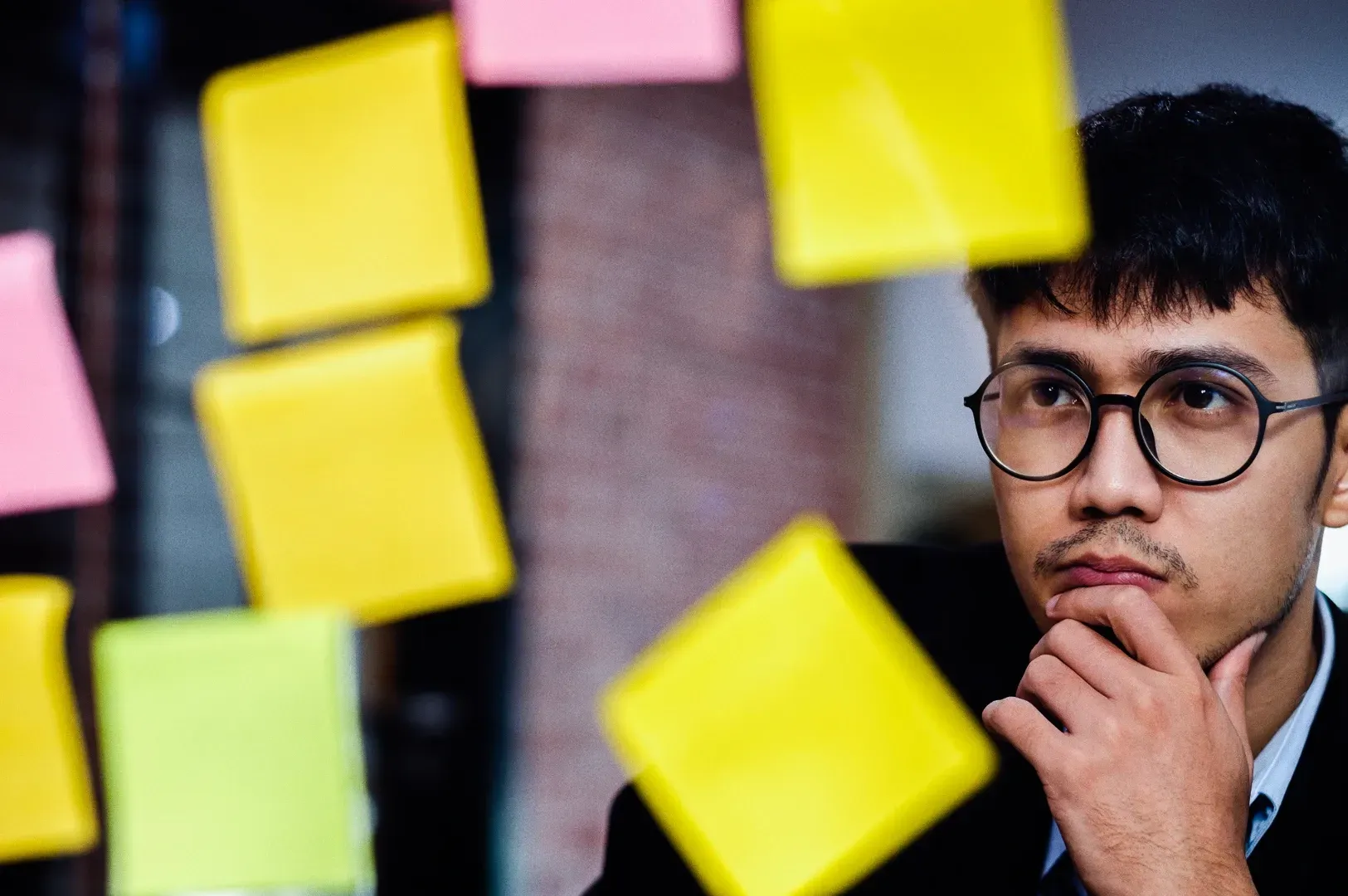 Eye-level medium close-up shot of a person seated behind a glass wall covered with colorful sticky notes, showing a workspace associated with planning, concentration, and focused thinking.