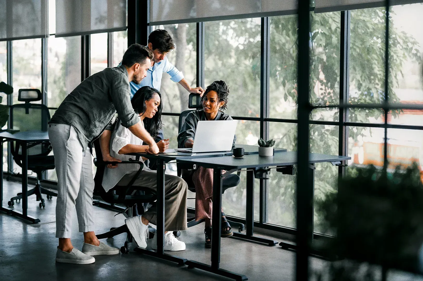 Eye-level medium-wide shot of a small group of professionals collaborating around a laptop at a table inside a modern office with large glass windows.