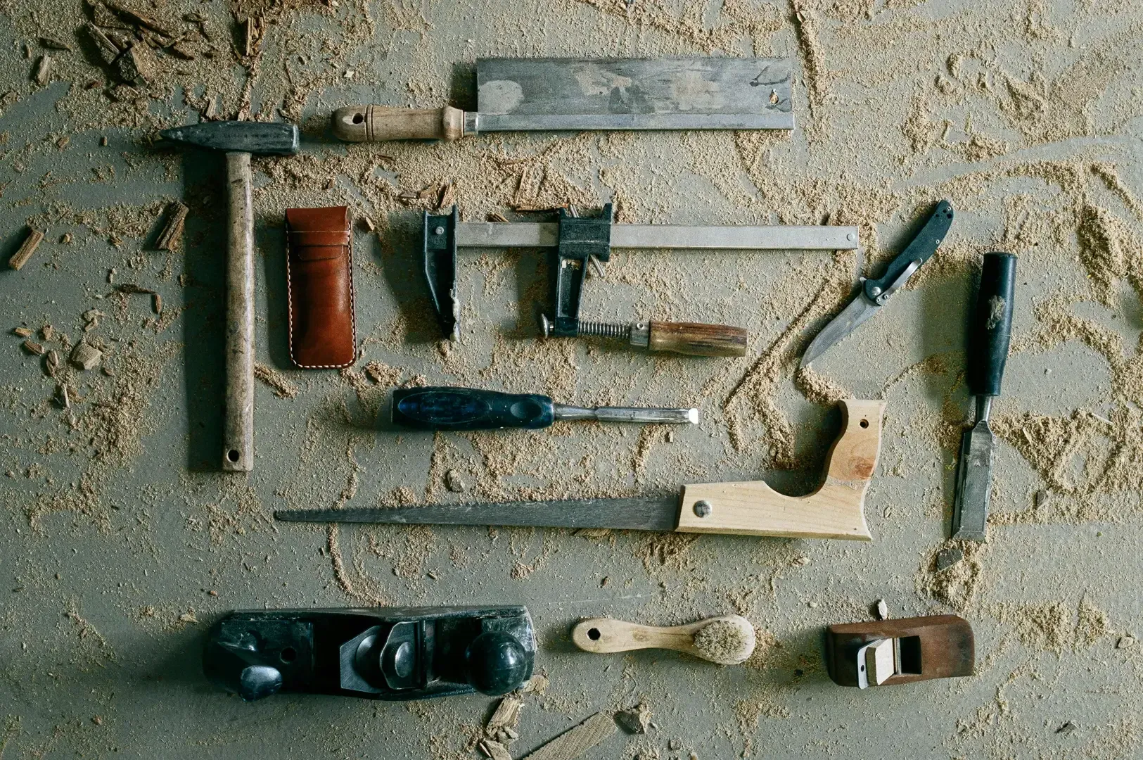 Top-down flat lay of essential woodworking hand tools on a sawdust-covered surface, including clamps, saws, chisels, and planer, captured in an overhead shot.