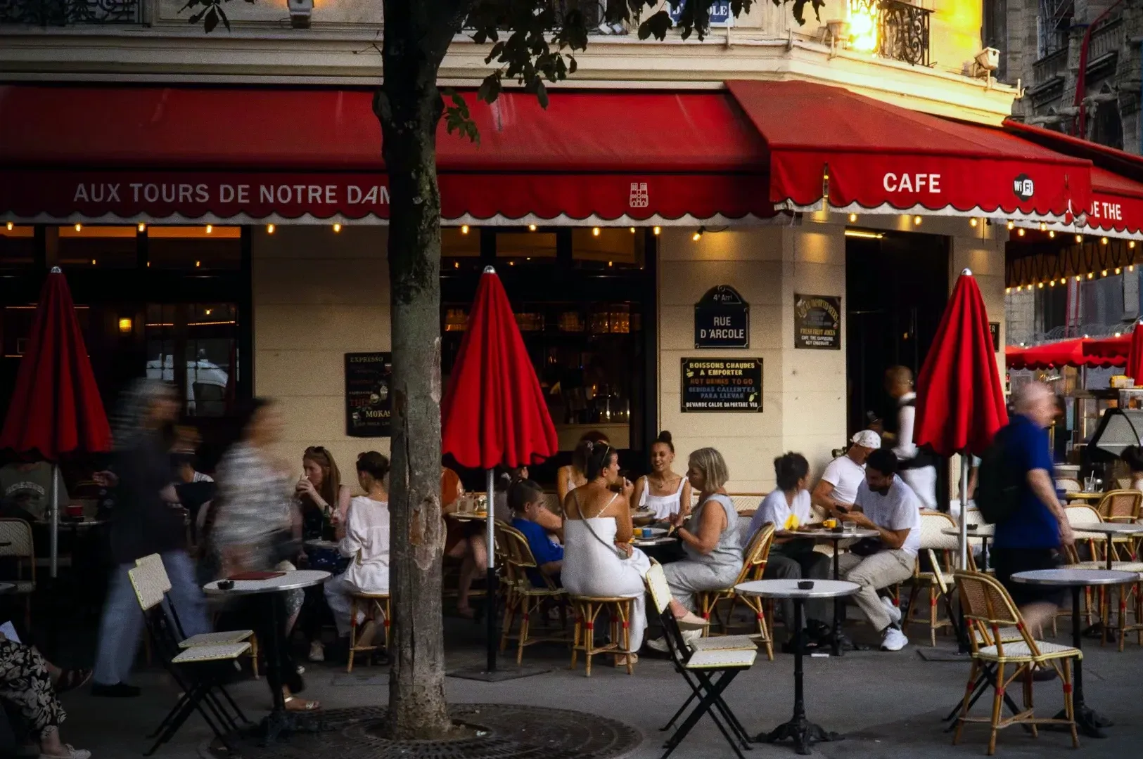 Bustling Paris street café with red awnings and outdoor seating, people gathered at small tables along the sidewalk, photographed from a street‑level wide angle, illustrating urban café culture and social “third places.”
