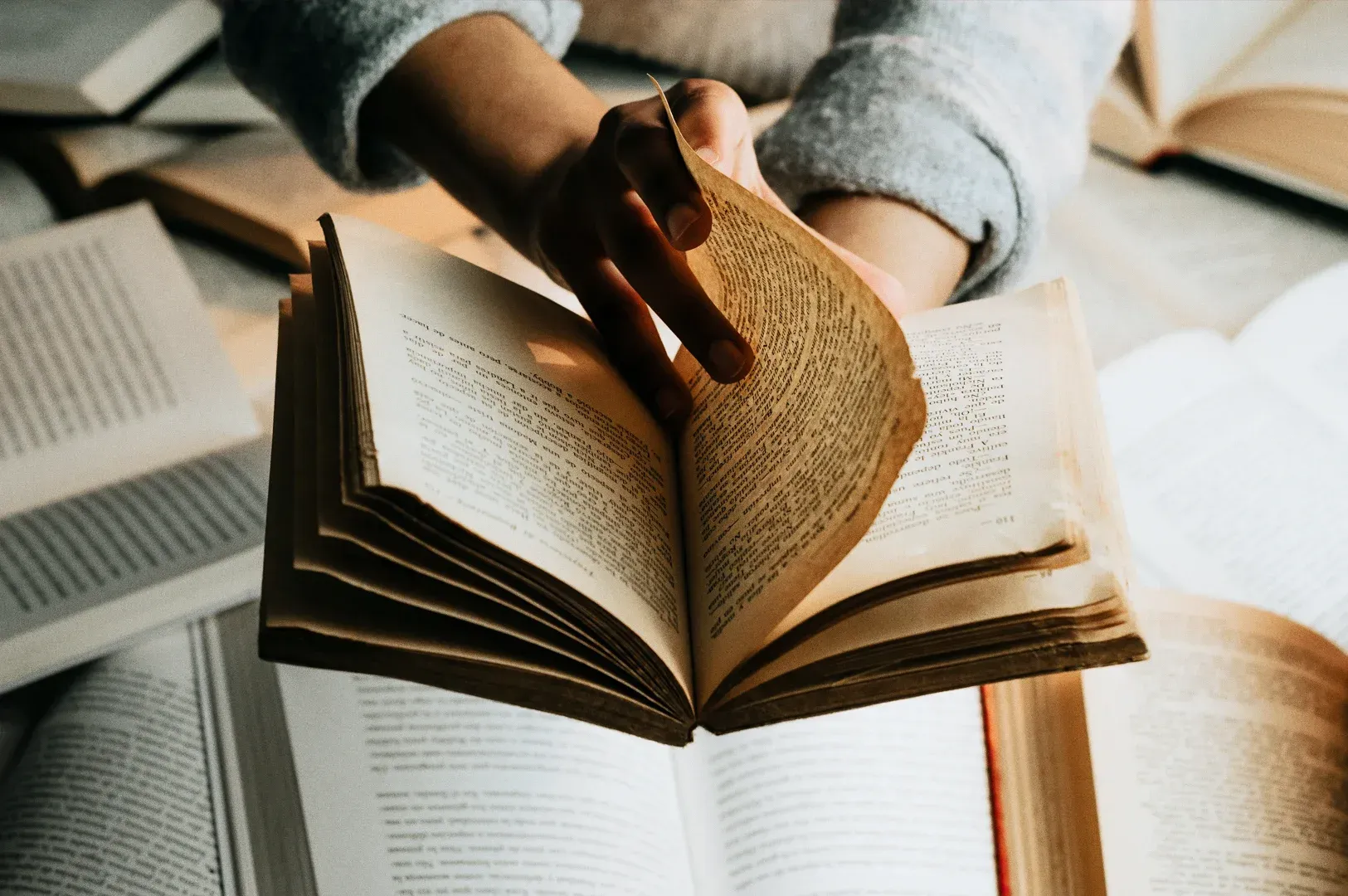 Close-up, slightly overhead shot of hands turning the pages of an open book placed among other books on a table, highlighting focused reading, learning, and knowledge study in a calm indoor setting.