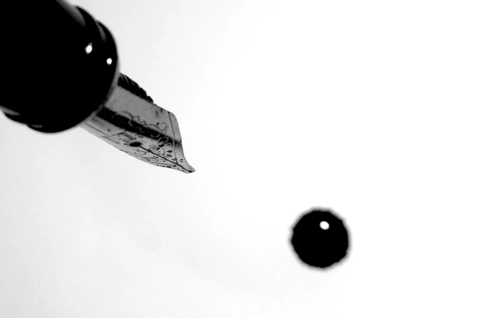Extreme macro top-down oblique shot of a fountain pen nib releasing a droplet of ink onto a blank surface, captured in high-contrast black and white.