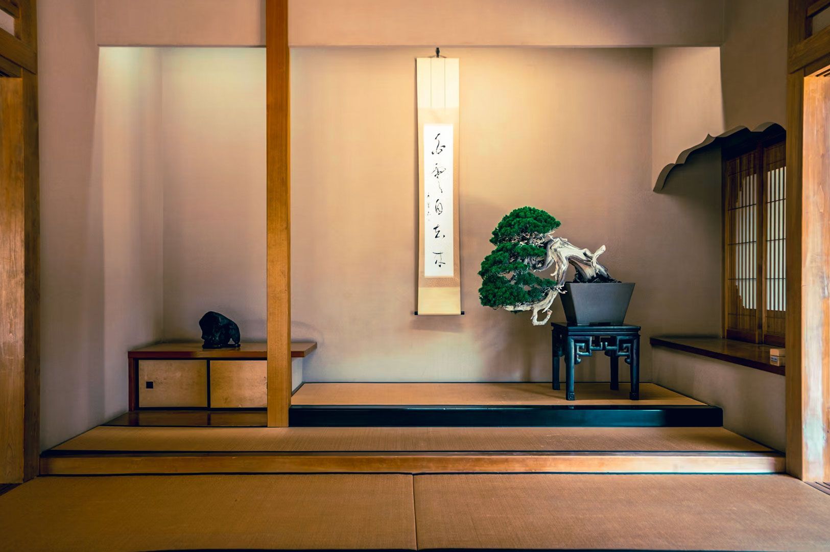 Minimalist Japanese room featuring tatami flooring, a bonsai display on a wooden stand, and a hanging calligraphy scroll.