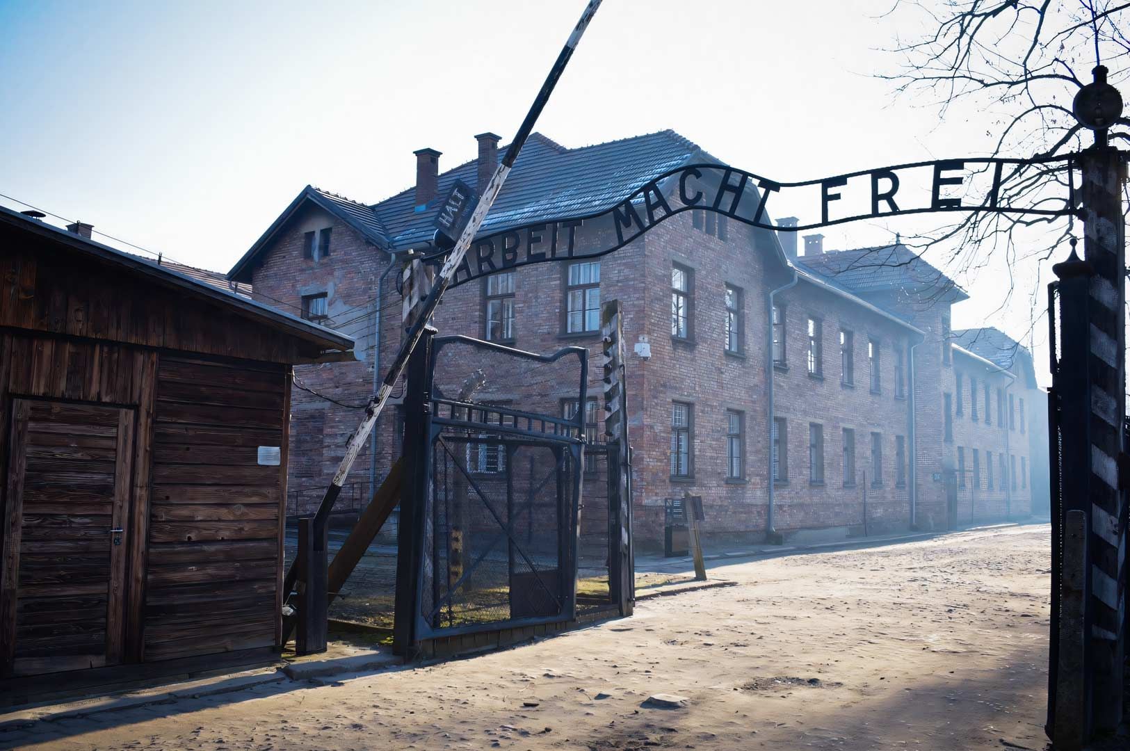 Entrance gate of the Auschwitz concentration camp with the historic ‘Arbeit macht frei’ sign above the walkway, showing brick barracks and wooden structures on a foggy day.