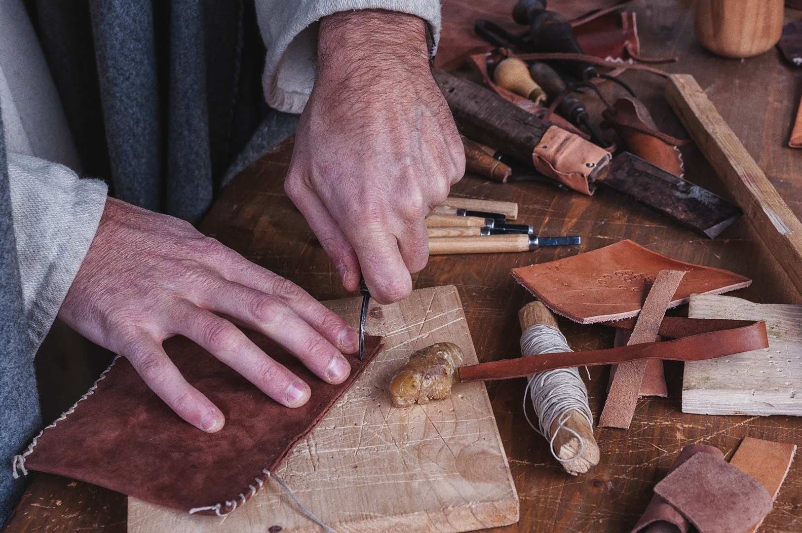 Close-up of hands working on brown leather with cutting tool, surrounded by thread, tools, and strips of leather.