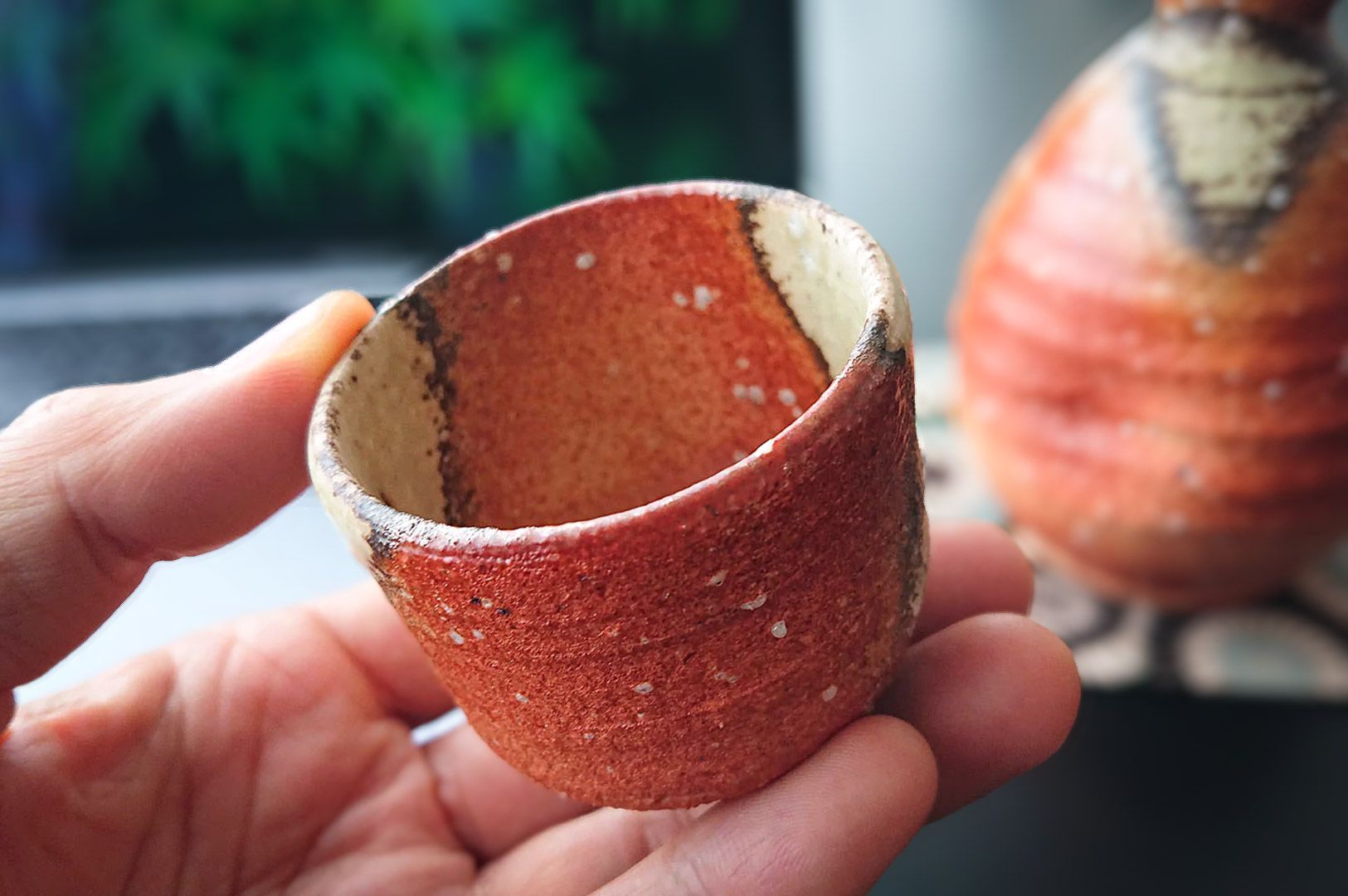 Hand holding a small handcrafted ceramic cup with a textured reddish‑brown glaze and artisanal design.