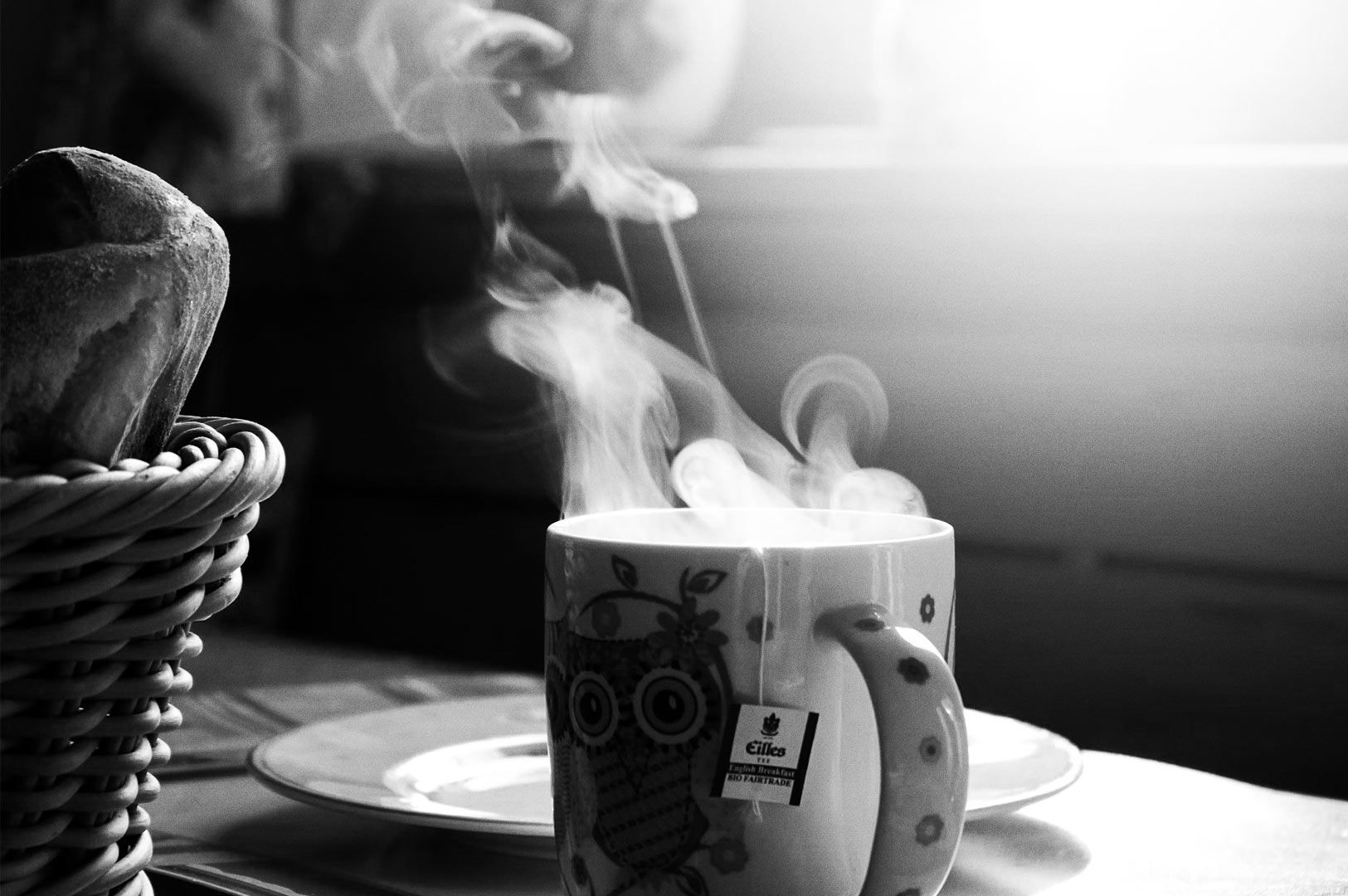 Steaming cup of tea in an owl‑patterned mug with a tea bag tag beside a wicker basket of bread in soft morning light.