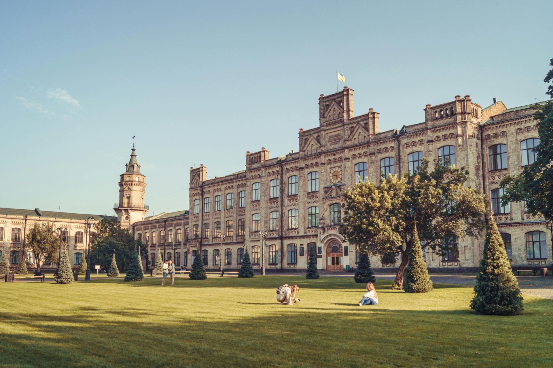 This image features a large, historic brick building with intricate architectural details and a central clock tower topped by a flag. People are scattered across a spacious green lawn in front of the structure, enjoying the sunny day amidst manicured conical trees.