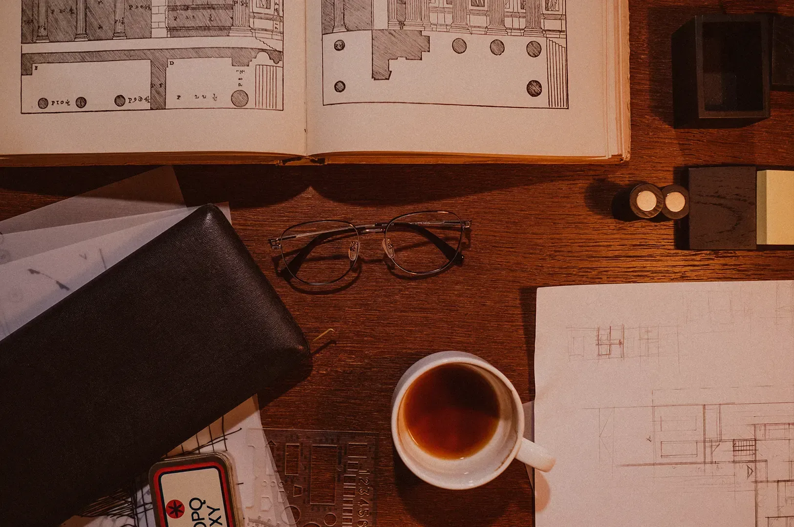 A top-down view of a wooden desk setup for creative work, featuring an open book with architectural diagrams, eyeglasses, a cup of coffee, and drafting tools, representing the essential morning rituals that foster focus.