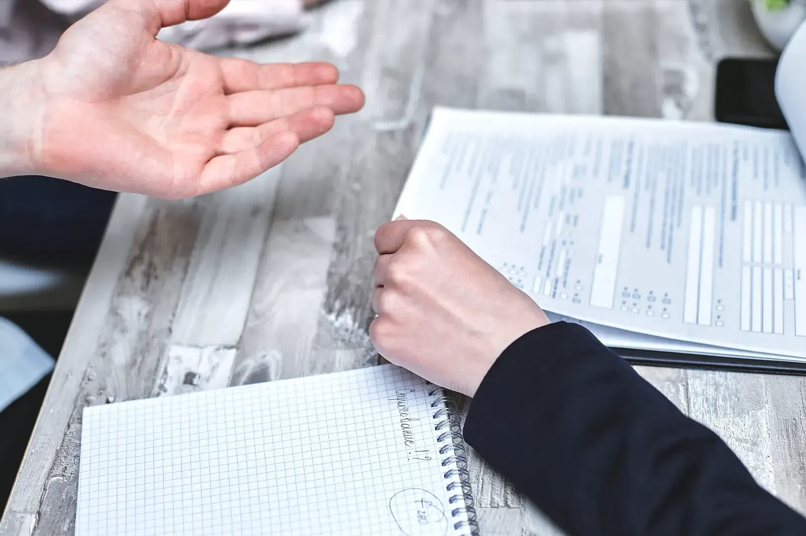 Two people collaborating over business paperwork and notes at a wooden desk, illustrating a conversation on envy as information rather than failure.