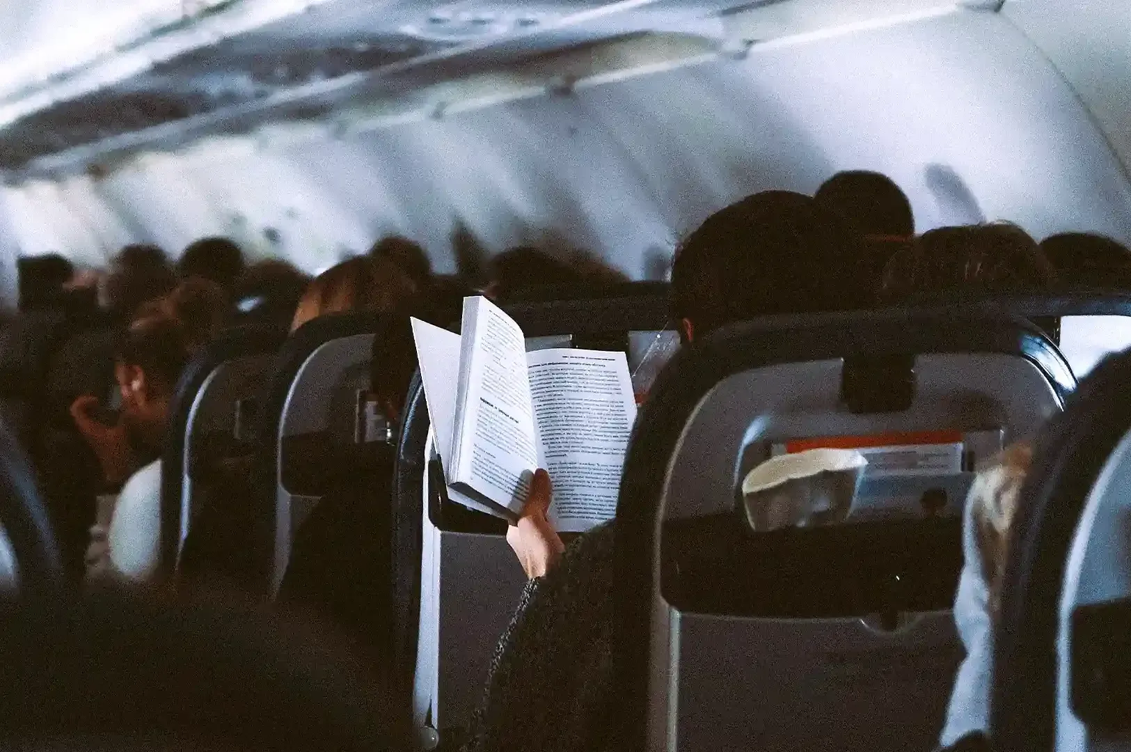 A passenger reading an open book mid-flight inside a dim airplane cabin, illustrating travel as a opportunity for reflection and seeing differently.