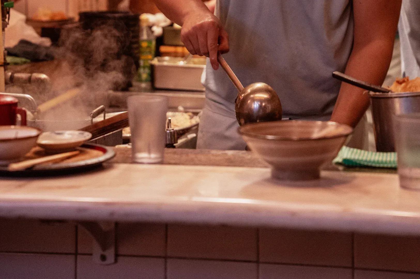 A close-up of a cook using a ladle in a steaming kitchen, highlighting the focused training and meticulous craft that ensure standards persist long after the original founder departs.