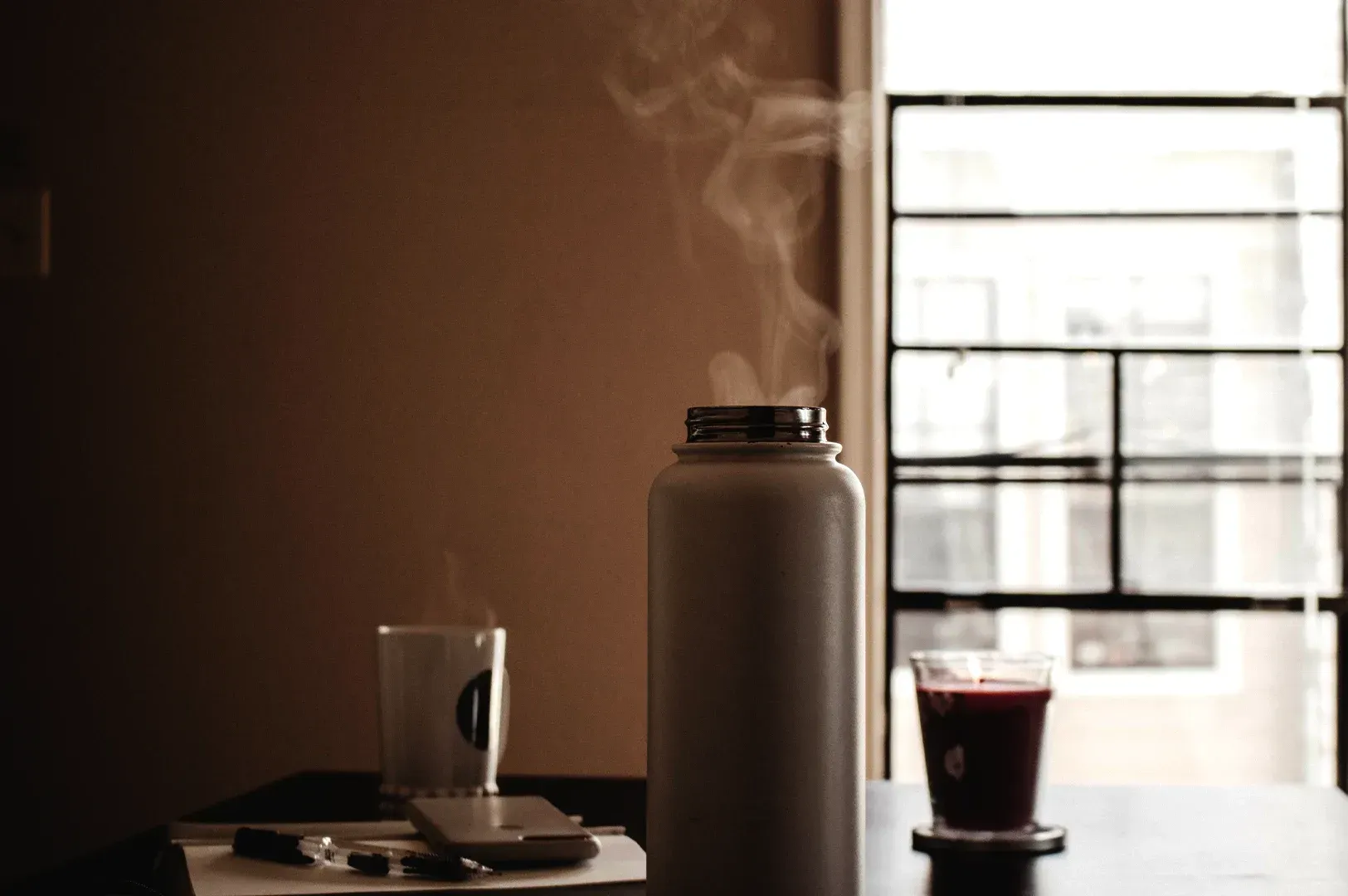 A steaming thermal bottle, a cup of tea, and notes on a quiet desk near a window, representing the atmosphere of deep focus, discipline, and the pursuit of excellence in a world that often ignores intangible values