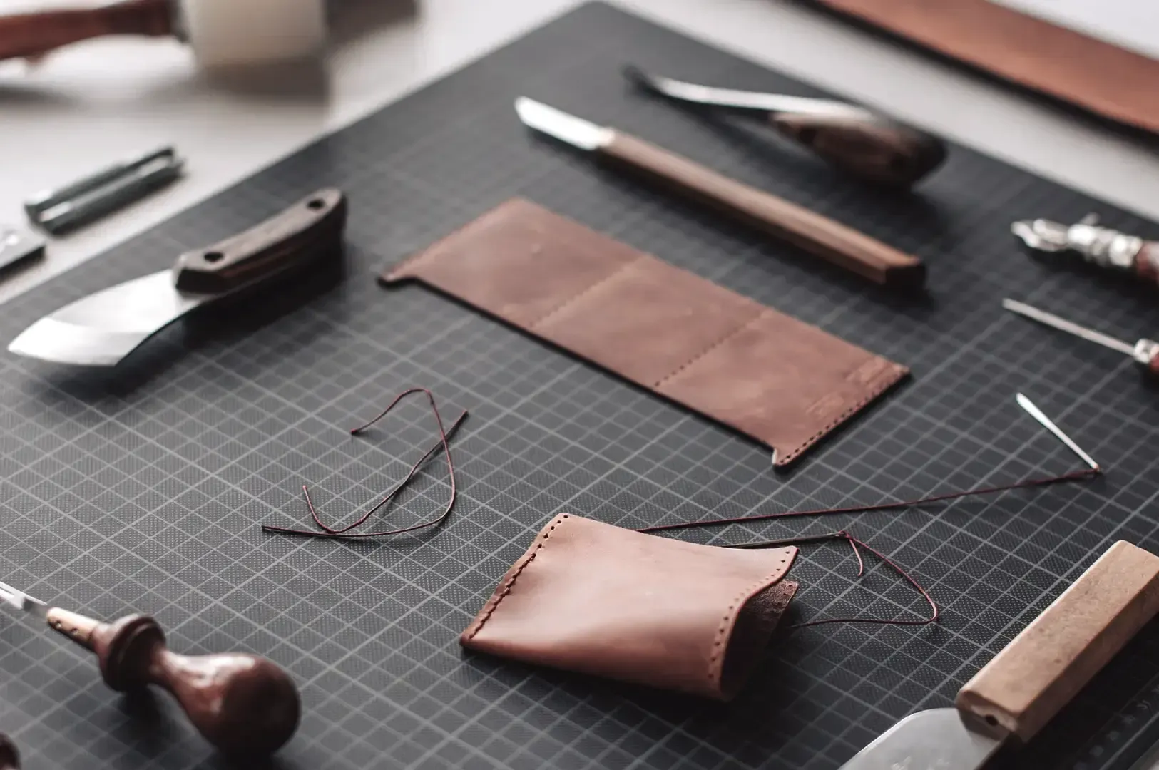An organized workstation featuring hand-stitched leather cardholders, needles, thread, and specialized cutting knives neatly arranged on a gridded mat.