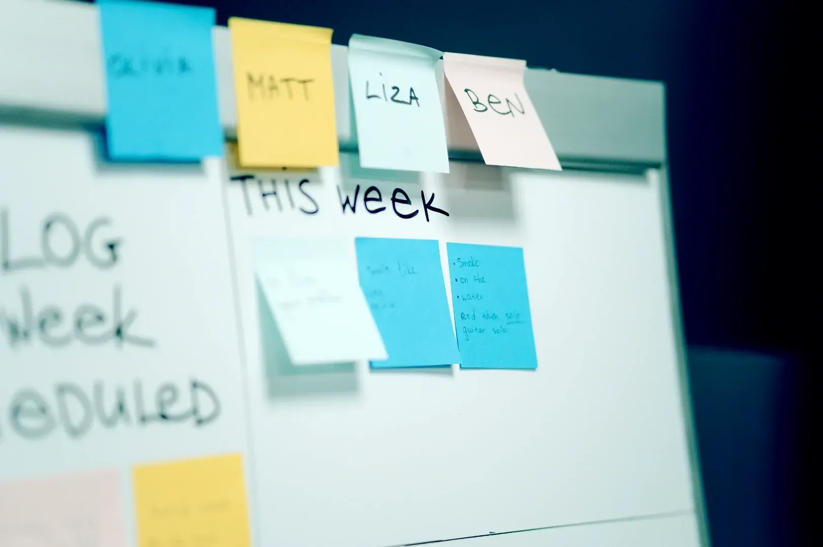 Whiteboard agile planning board filled with colorful sticky notes labeled with team member names, illustrating daily task tracking and team accountability.
