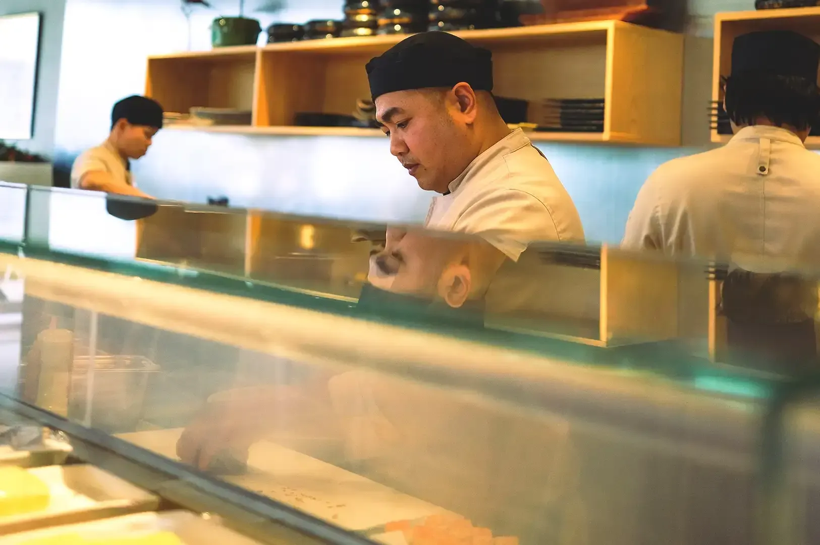 A focused sushi chef working meticulously behind a glass counter, embodying the philosophy of learning a craft deeply enough to bend it with care through stewardship rather than rebellion.