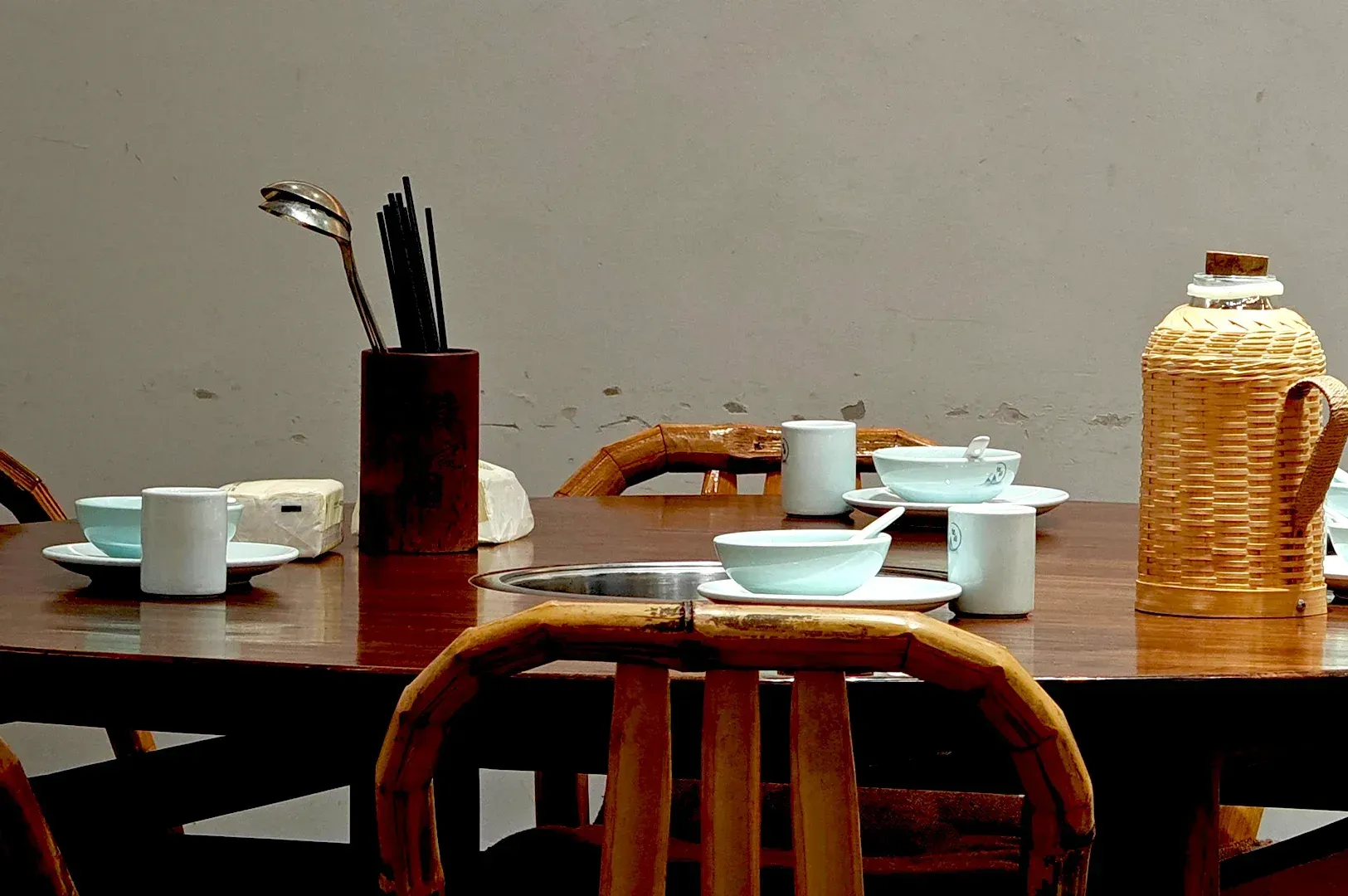 A traditional wooden dining table set with rustic ceramic bowls, bamboo chairs, and a woven tea thermos, highlighting how human-made, textured objects ground us in authenticity.