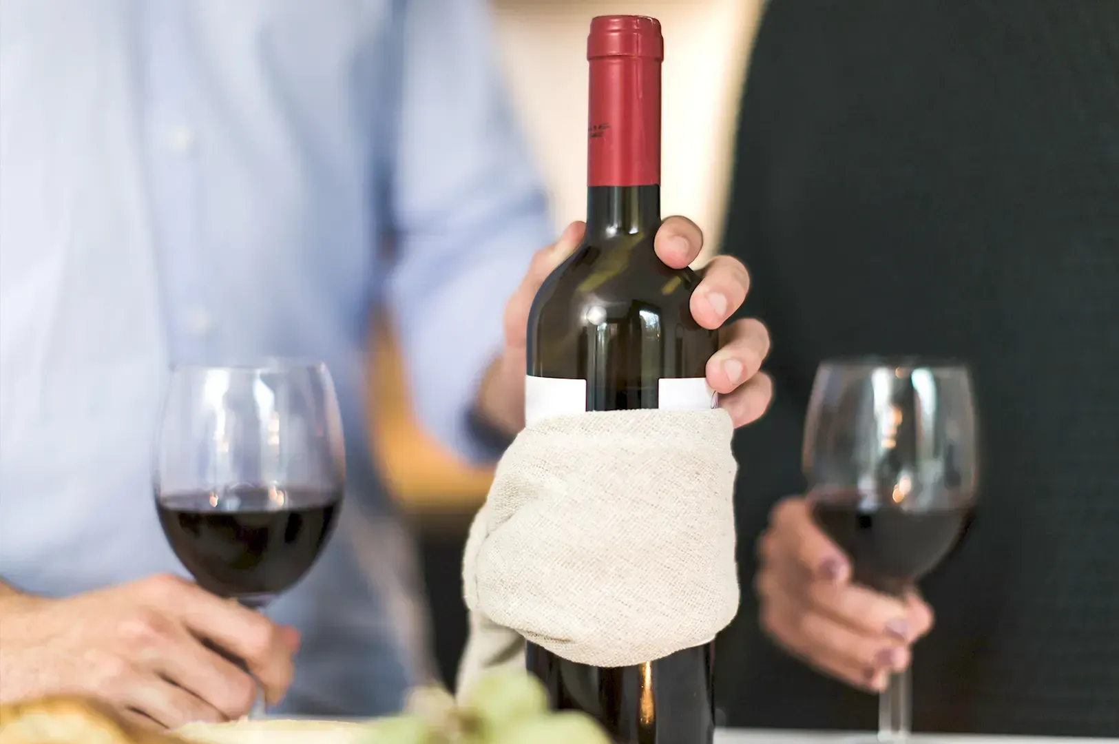 A person holding a bottle of red wine wrapped in a cloth napkin, flanked by two filled wine glasses, capturing a quiet moment of wine appreciation and taste.