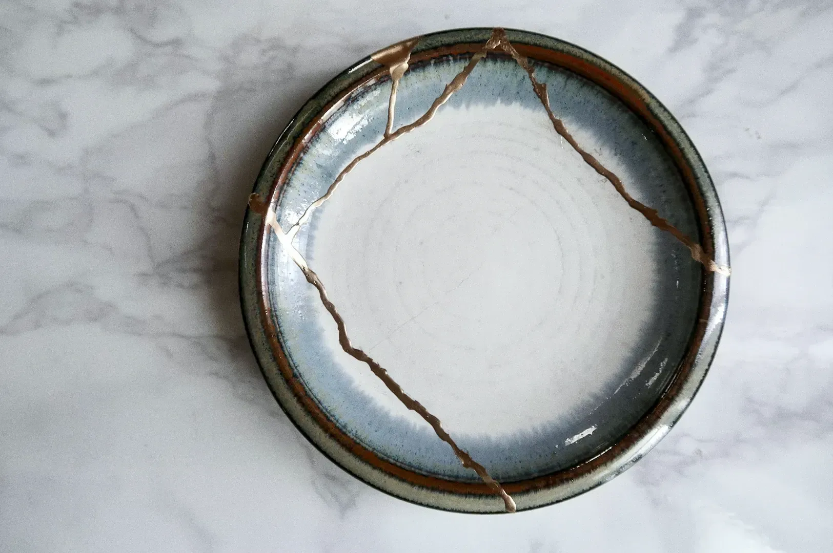 Top-down close-up shot of a repaired ceramic plate with visible kintsugi cracks on a marble surface, representing repair culture, durability, and the value of keeping items that last