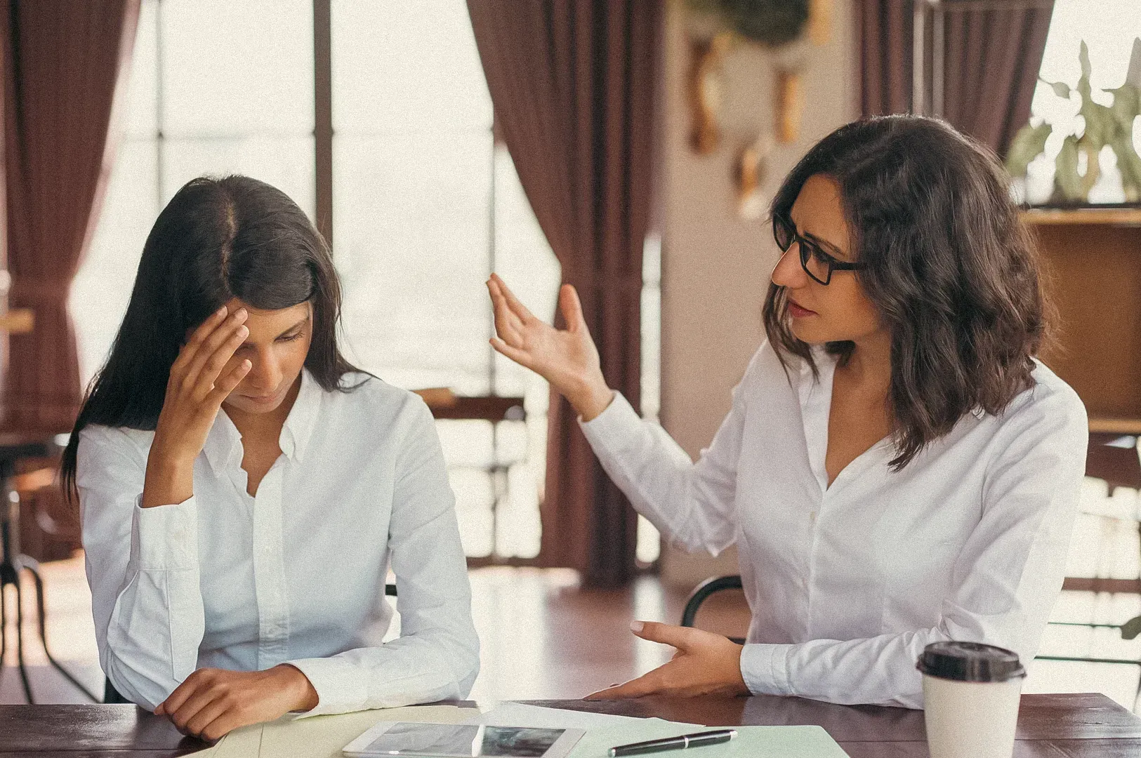 Two colleagues in a workplace setting engaged in a serious conversation at a desk with papers and a tablet.