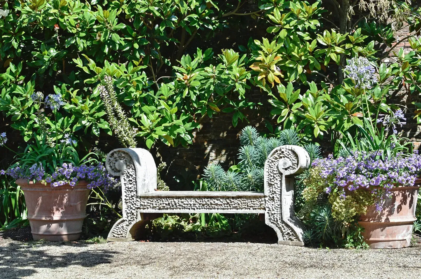 Eye-level wide shot of a carved stone bench surrounded by lush greenery and flowering plants in a quiet garden courtyard, illustrating slow travel, mindful observation, and curated peaceful outdoor experiences