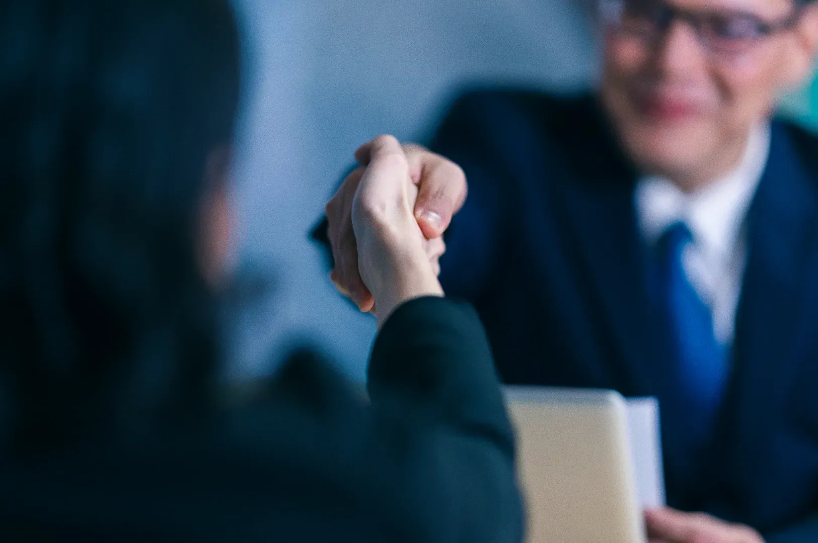 Shallow depth-of-field close-up shot of two professionals shaking hands across a desk in a formal business meeting setting.