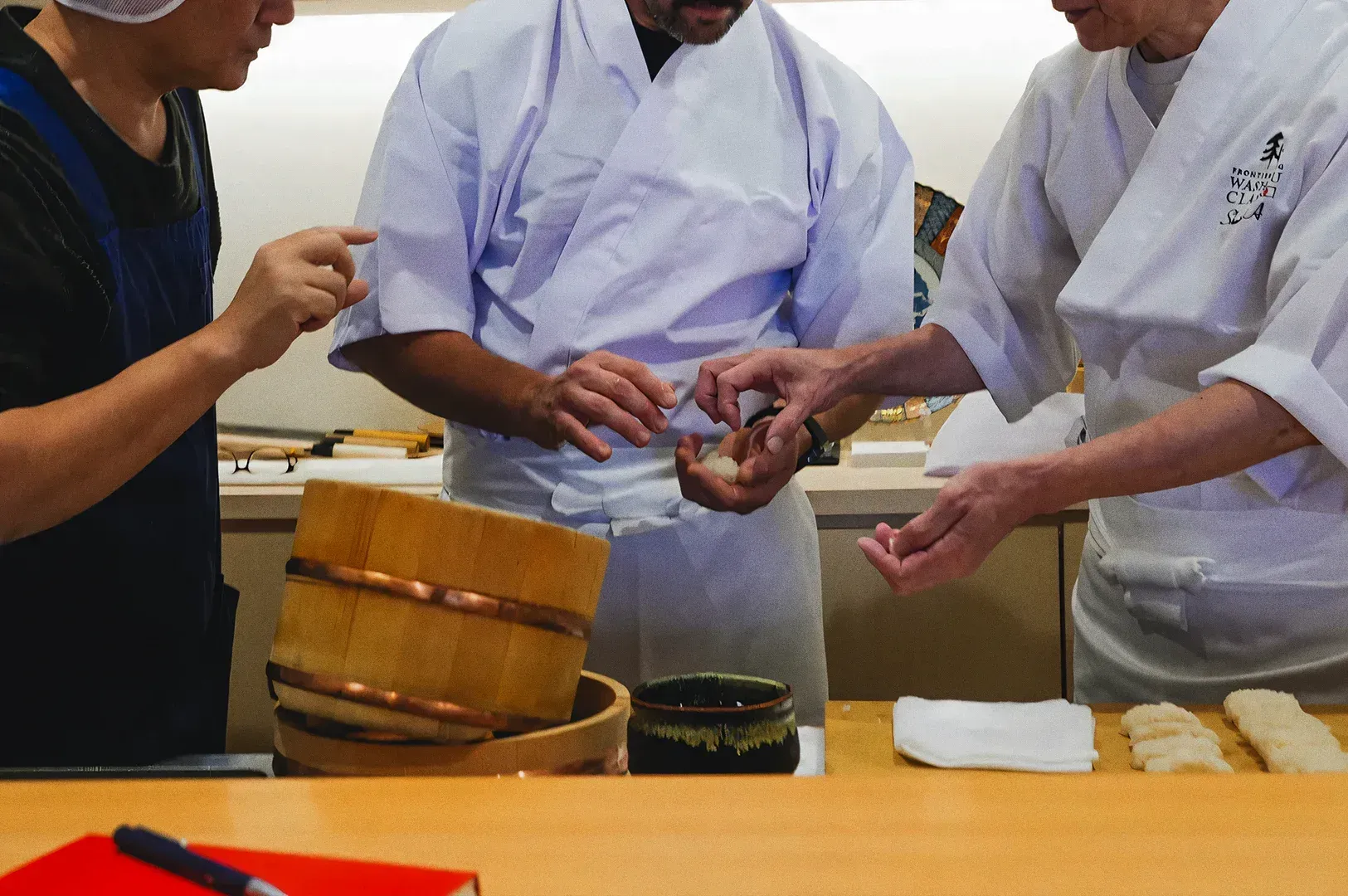 Close-up of sushi chefs shaping rice by hand at a traditional Japanese restaurant counter with wooden bucket and utensils, captured in a medium close-up shot at counter level.