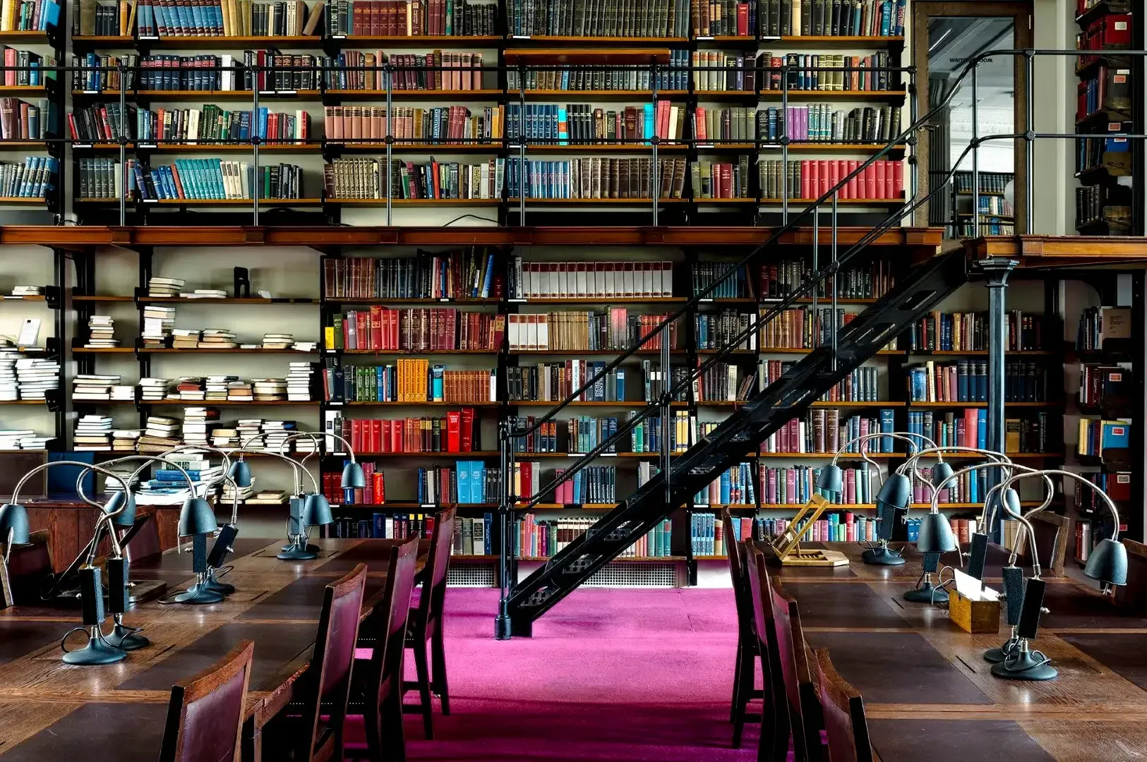 Expansive library interior featuring multi‑level bookshelves, a metal staircase, and long wooden reading tables with study lamps.