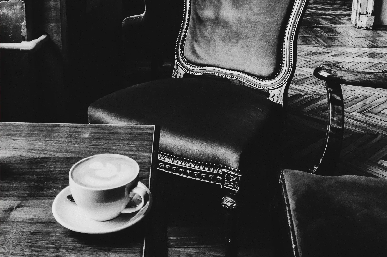 Black‑and‑white café interior with a cappuccino on a wooden table beside an empty upholstered chair with carved details, photographed from a seated eye‑level angle, evoking European coffeehouse culture and the concept of the “third place.”