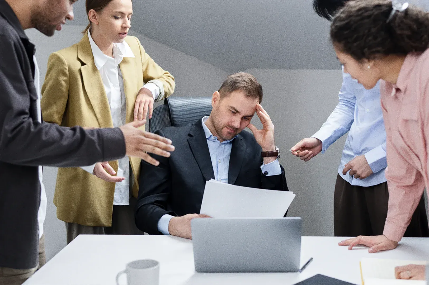 Medium-wide, eye-level shot of a workplace meeting where a professional sits at a desk reviewing documents on a laptop while colleagues stand nearby, illustrating teamwork, collaboration, and leadership decision-making in an office environment.