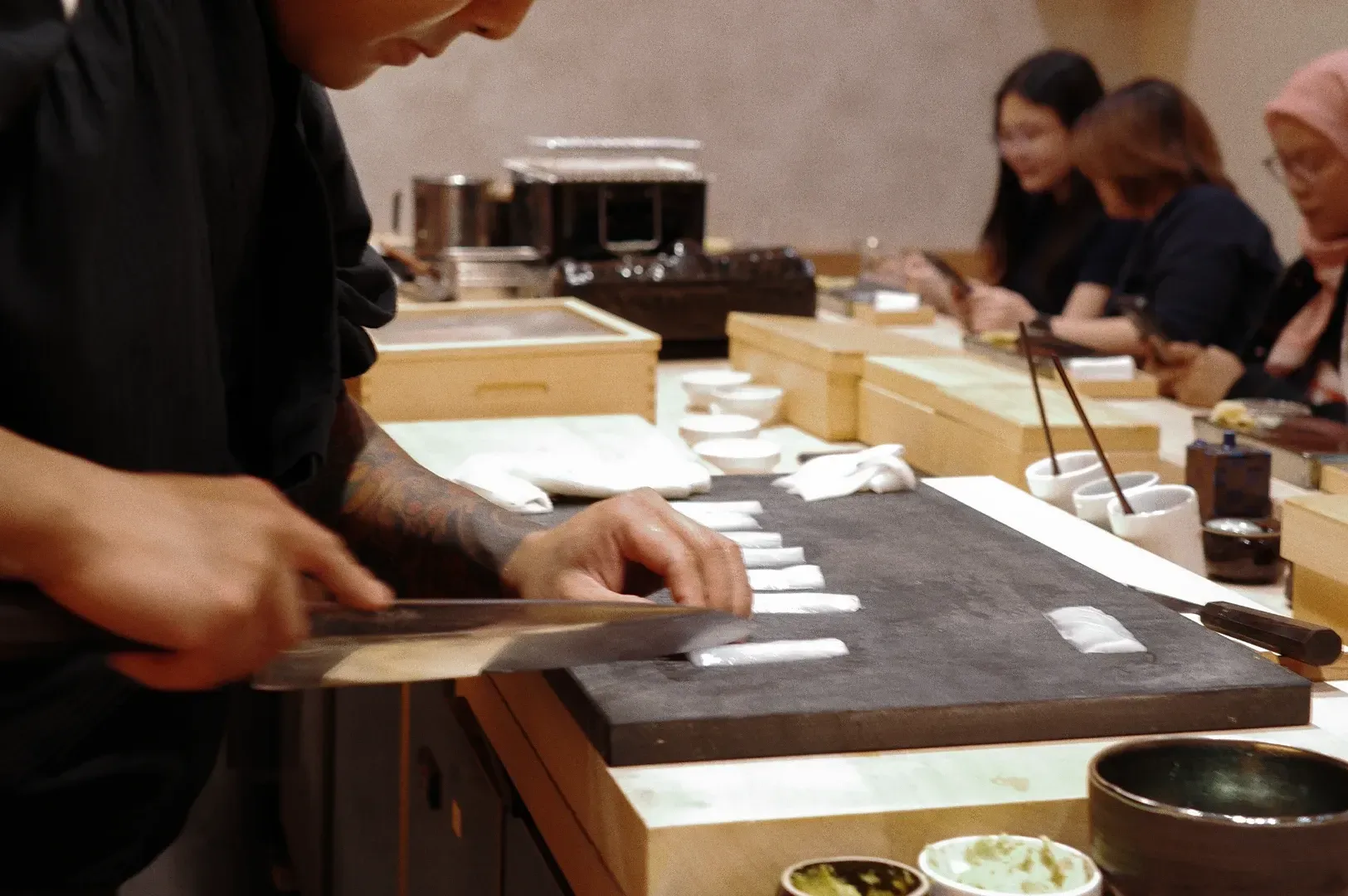 Close-up side-angle shot of a chef slicing fresh fish into uniform pieces on a stone board at a sushi counter, with seated diners softly blurred in the background of a minimalist Japanese restaurant.