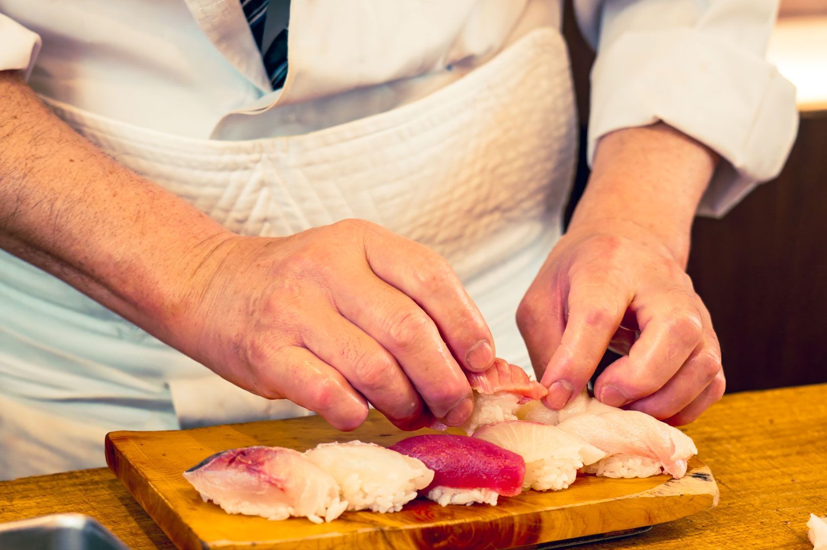 Chef’s hands carefully forming assorted nigiri sushi pieces on a wooden board in a traditional Japanese kitchen.