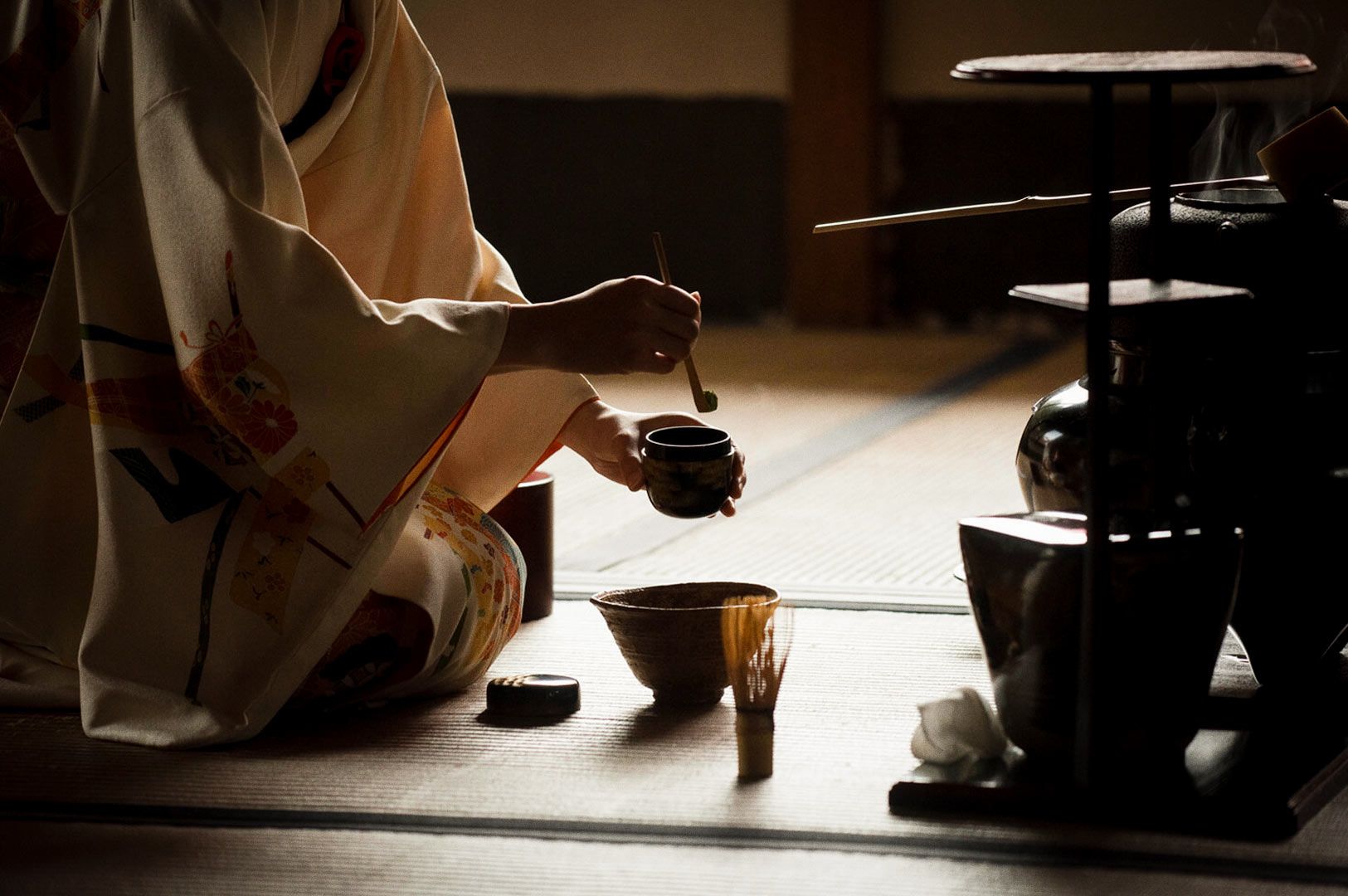 A traditional tea ceremony scene showing a person in ornate robes preparing tea utensils on a tatami mat, with soft lighting highlighting the ceremonial setup.